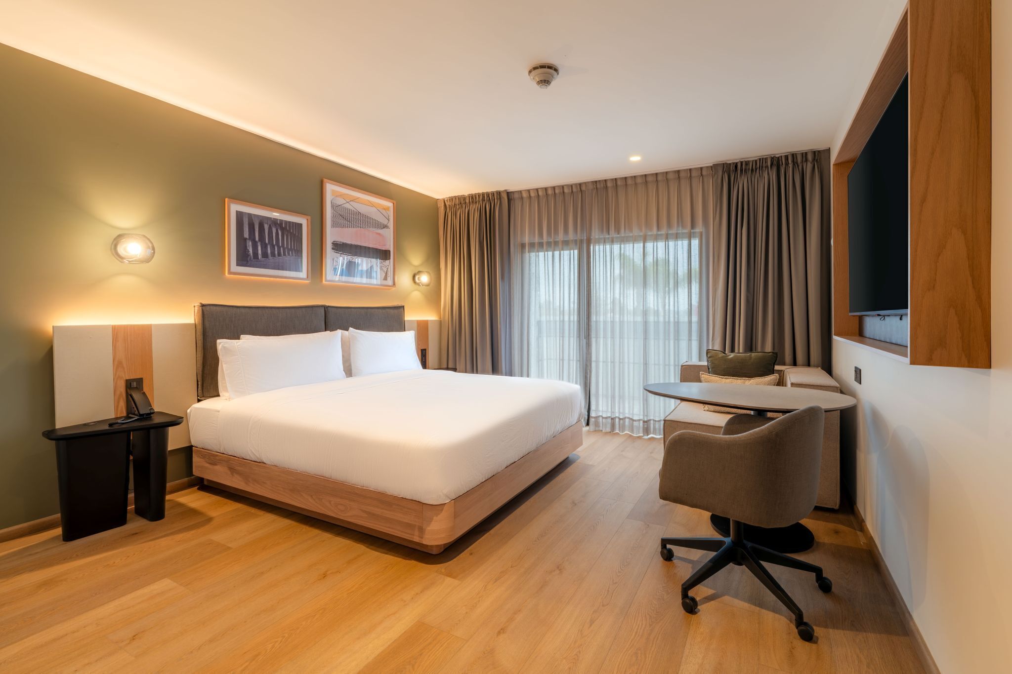 A hotel room with a bed, bedside table, lamp, desk with chair, and a TV mounted on the wall.