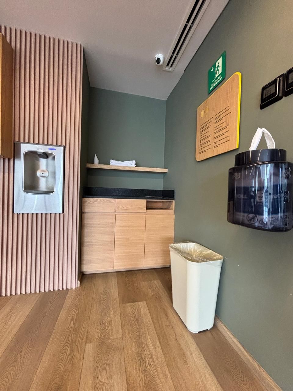 Habitación de hotel con piso de madera, aire acondicionado de pared, dispensador de agua, basurero y dispensador de bolsas de pared.
