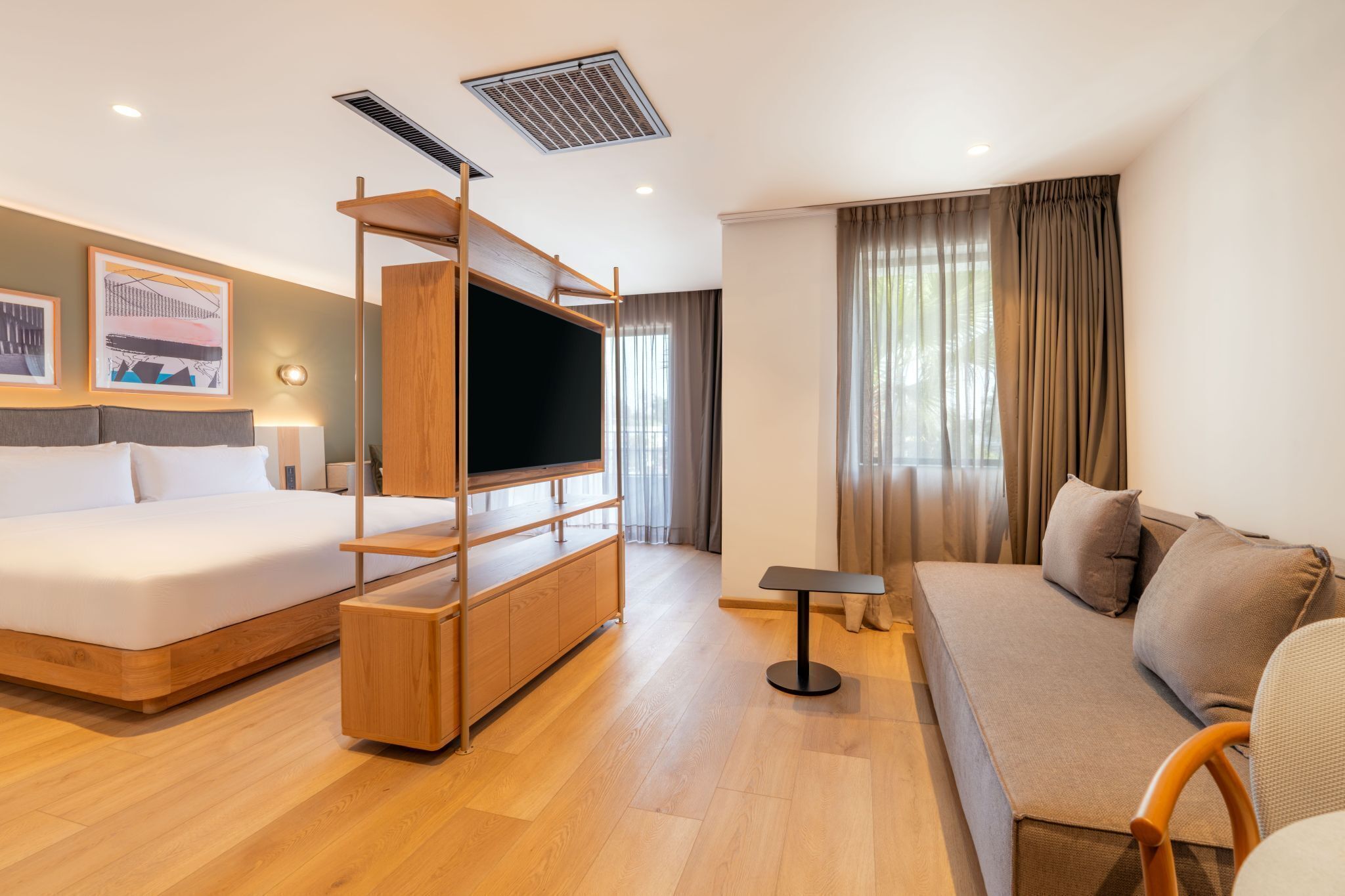 Quarto de hotel moderno com cama, sofá, televisão sobre um suporte de madeira e uma mesinha.