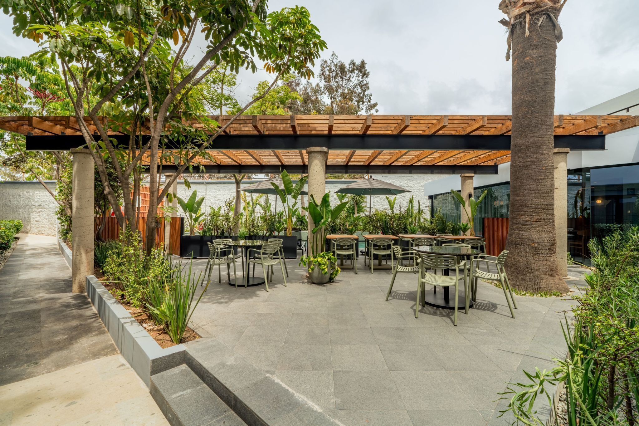 An outdoor patio area with multiple tables and chairs, surrounded by greenery and a wooden pergola.