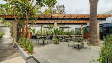 An outdoor patio area with multiple tables and chairs, surrounded by greenery and a wooden pergola.