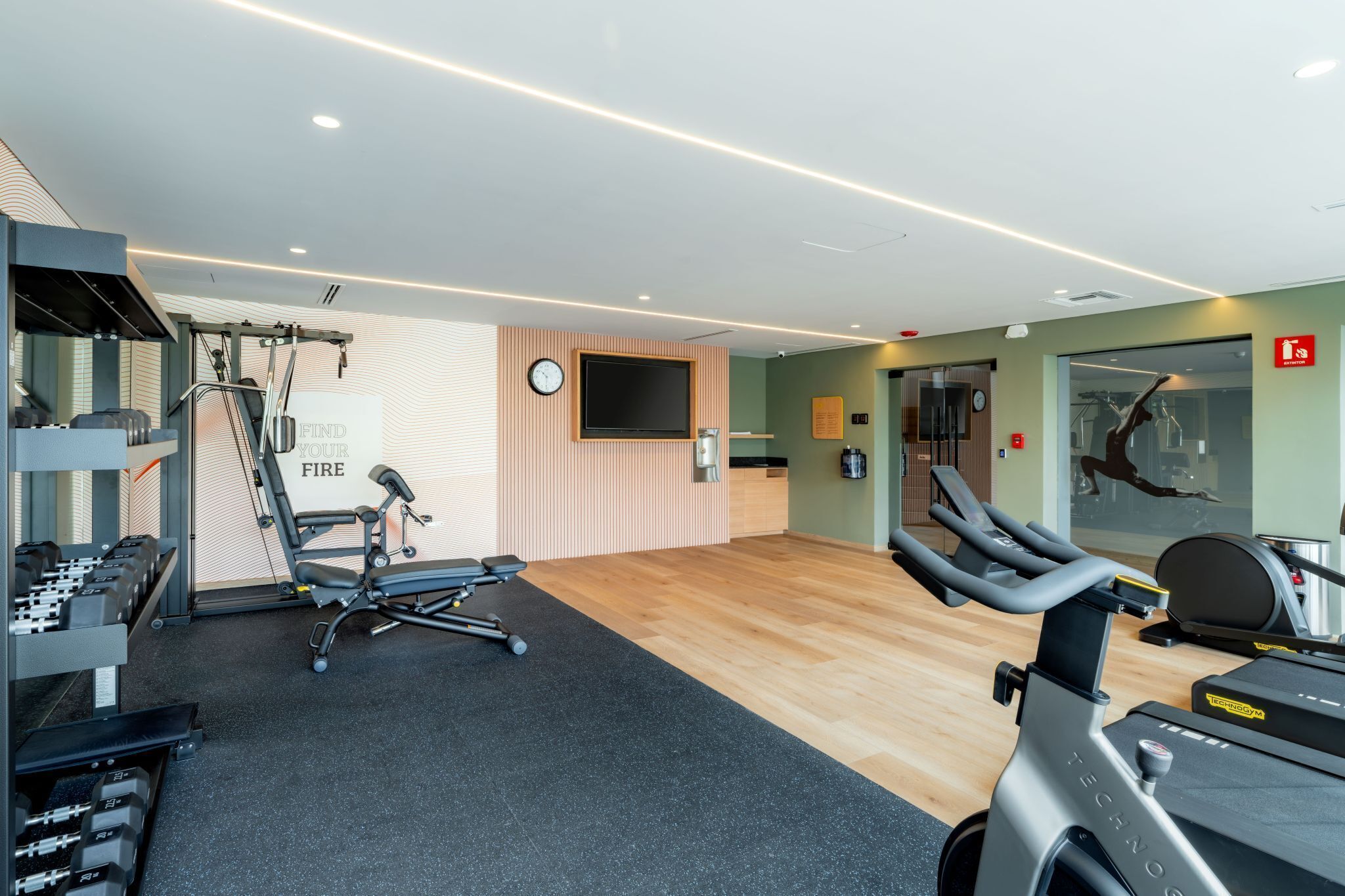 Uma academia com diversos equipamentos de exercício, uma televisão presa à parede e um espelho.