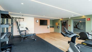 Uma academia com diversos equipamentos de exercício, uma televisão presa à parede e um espelho.