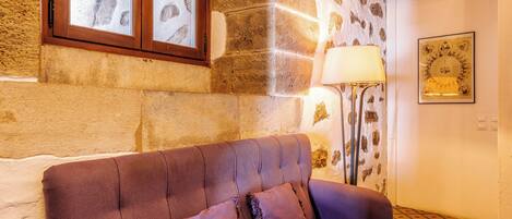 A room with a brown leather sofa, a stone wall, a window with wooden shutters, a floor lamp, and a framed picture on the wall.