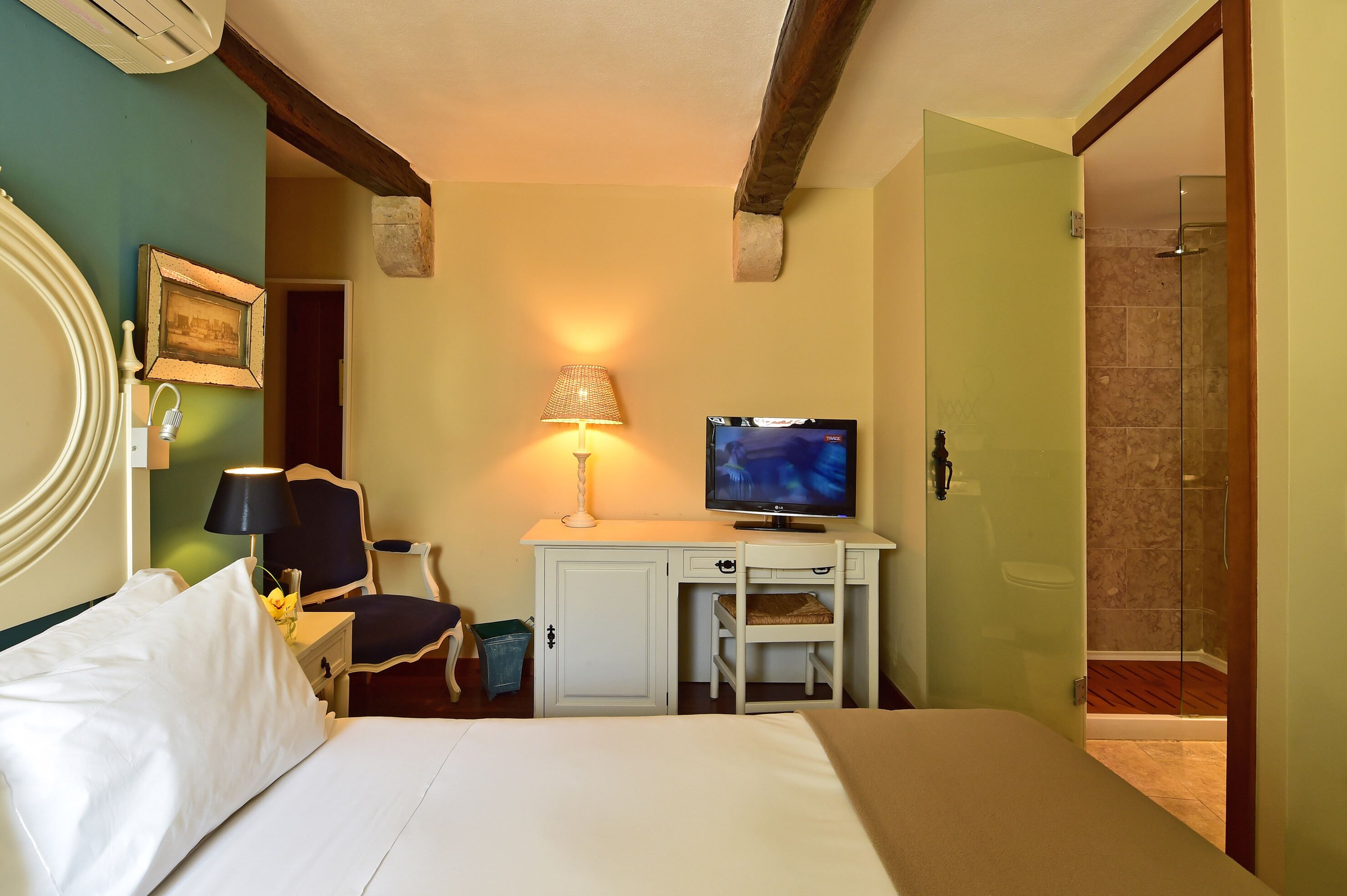 Habitación de hotel con cama, escritorio con televisor, una silla, lámpara y zona de ducha.