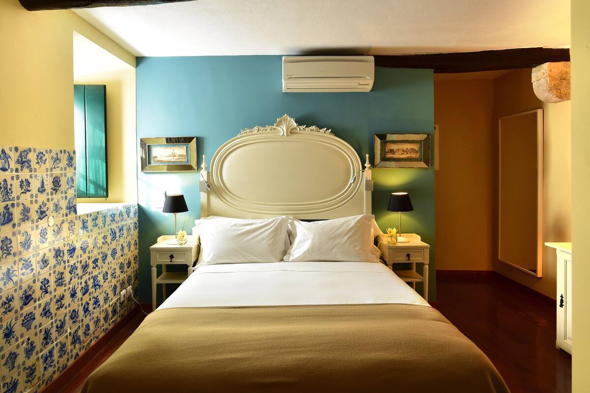 Ein Hotelzimmer mit einem großen Bett, zwei Nachttischen mit Lampen, einer Wand-Klimaanlage und einer Wand mit Kachelfliesen in floralem Muster.