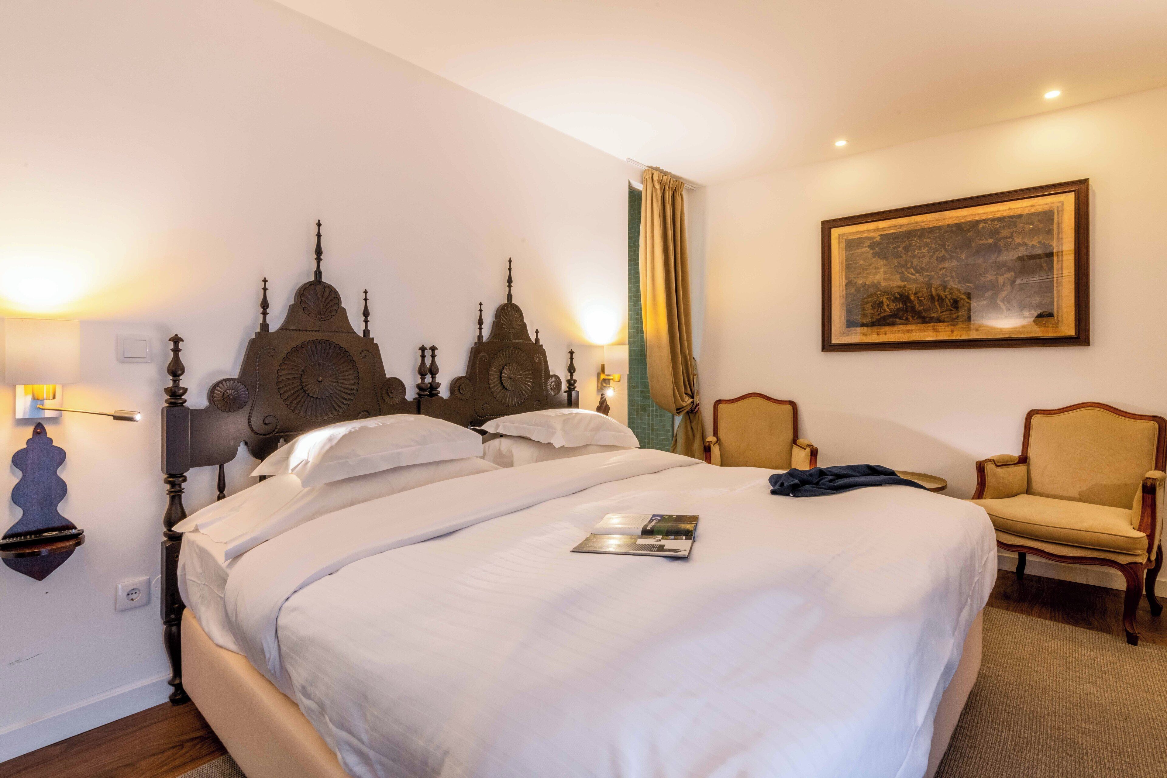 A hotel room with a large bed, two chairs, a painting on the wall, and a lamp.