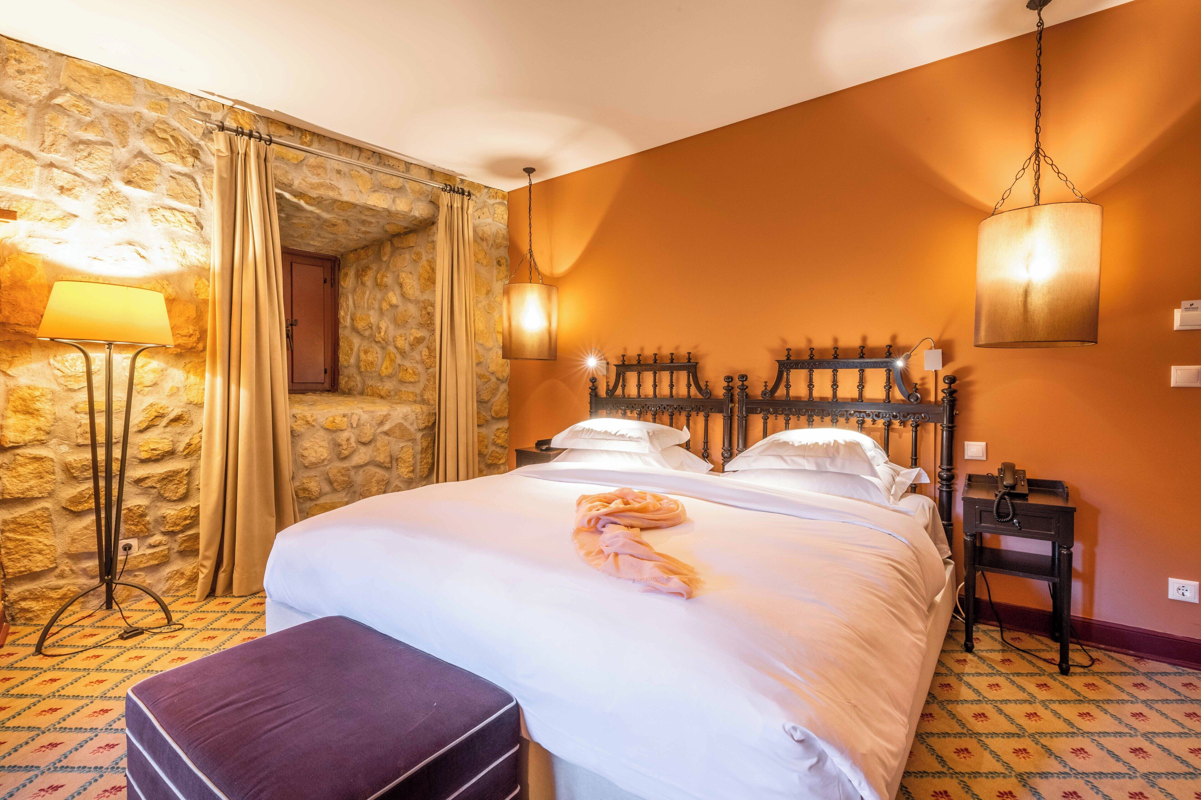 Habitación de hotel con pared de piedra, cama con ropa blanca, un banco morado y una lámpara colgante.