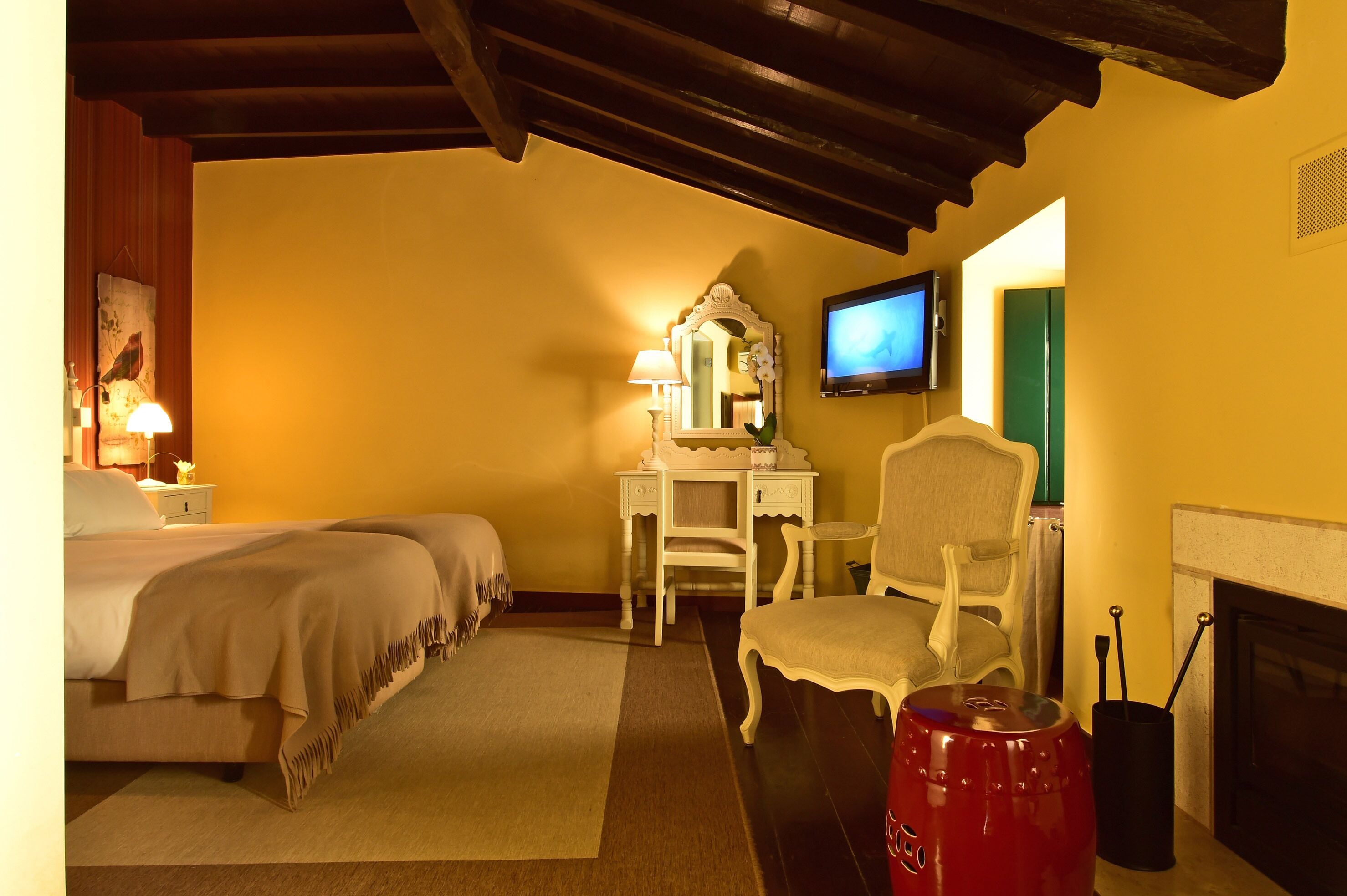 A hotel room with two beds, a desk with a mirror, a television, a fireplace, and a red kettle.