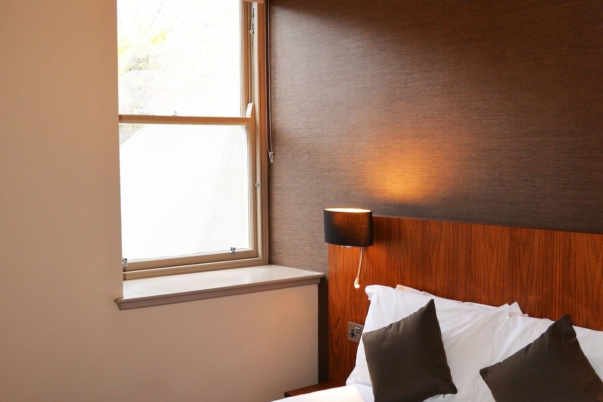 Quarto de hotel com uma cama, janela e luminária fixada na parede.