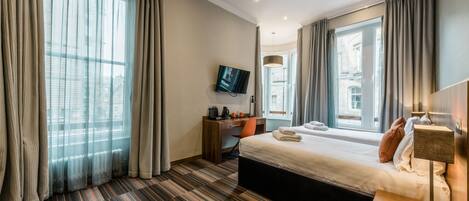 A modern hotel room with a large bed, a desk with a chair, a flat-screen TV, and large windows with curtains.