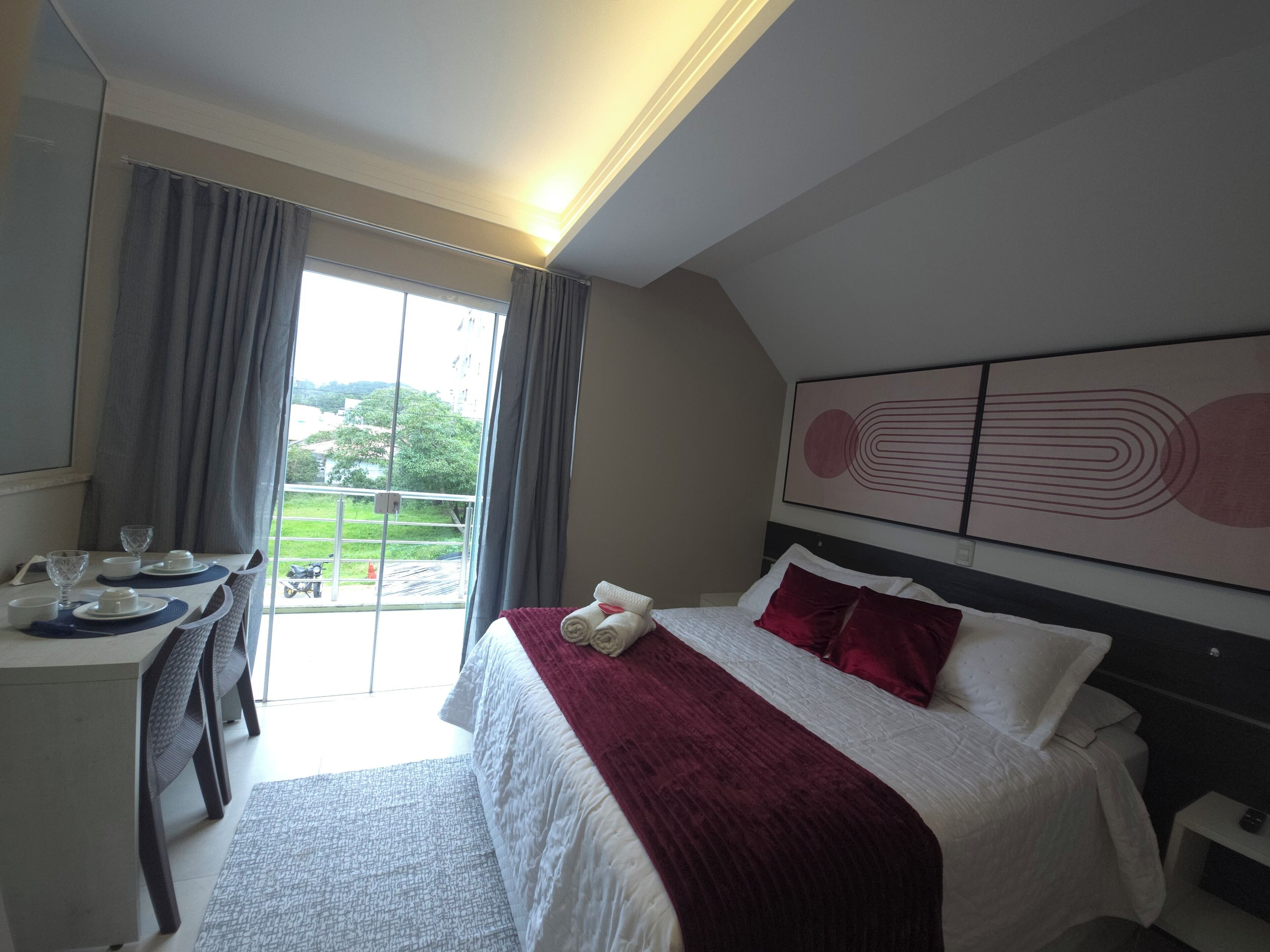 A hotel room with a bed, a desk with a chair, a window with curtains, and a painting on the wall.