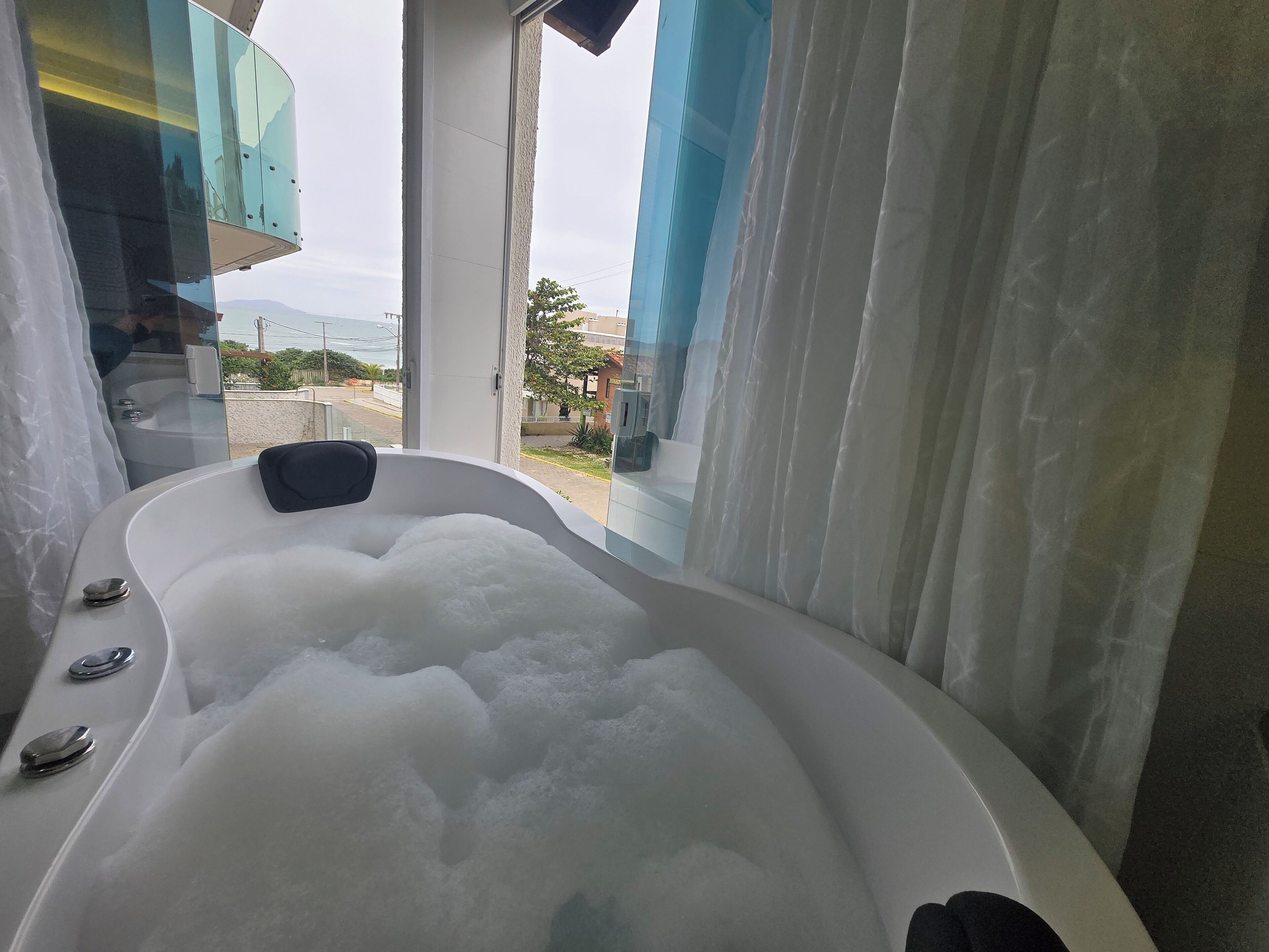 A modern bathroom with a large bathtub filled with bubbles, a glass shower enclosure, and a view of the outdoors.