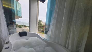 A modern bathroom with a large bathtub filled with bubbles, a glass shower enclosure, and a view of the outdoors.