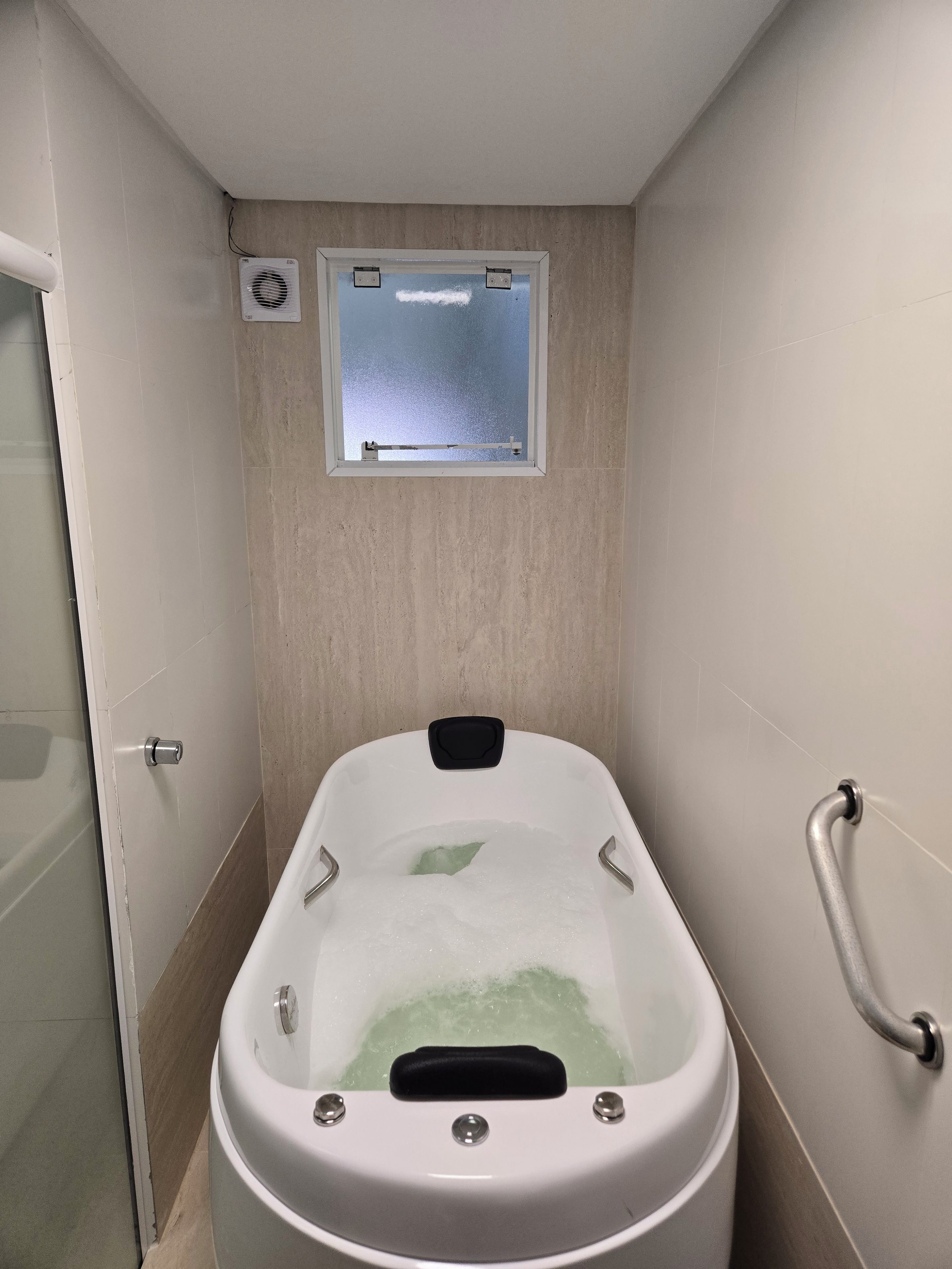 A bathtub with a window above it, a shower door to the left, and a grab bar on the right.