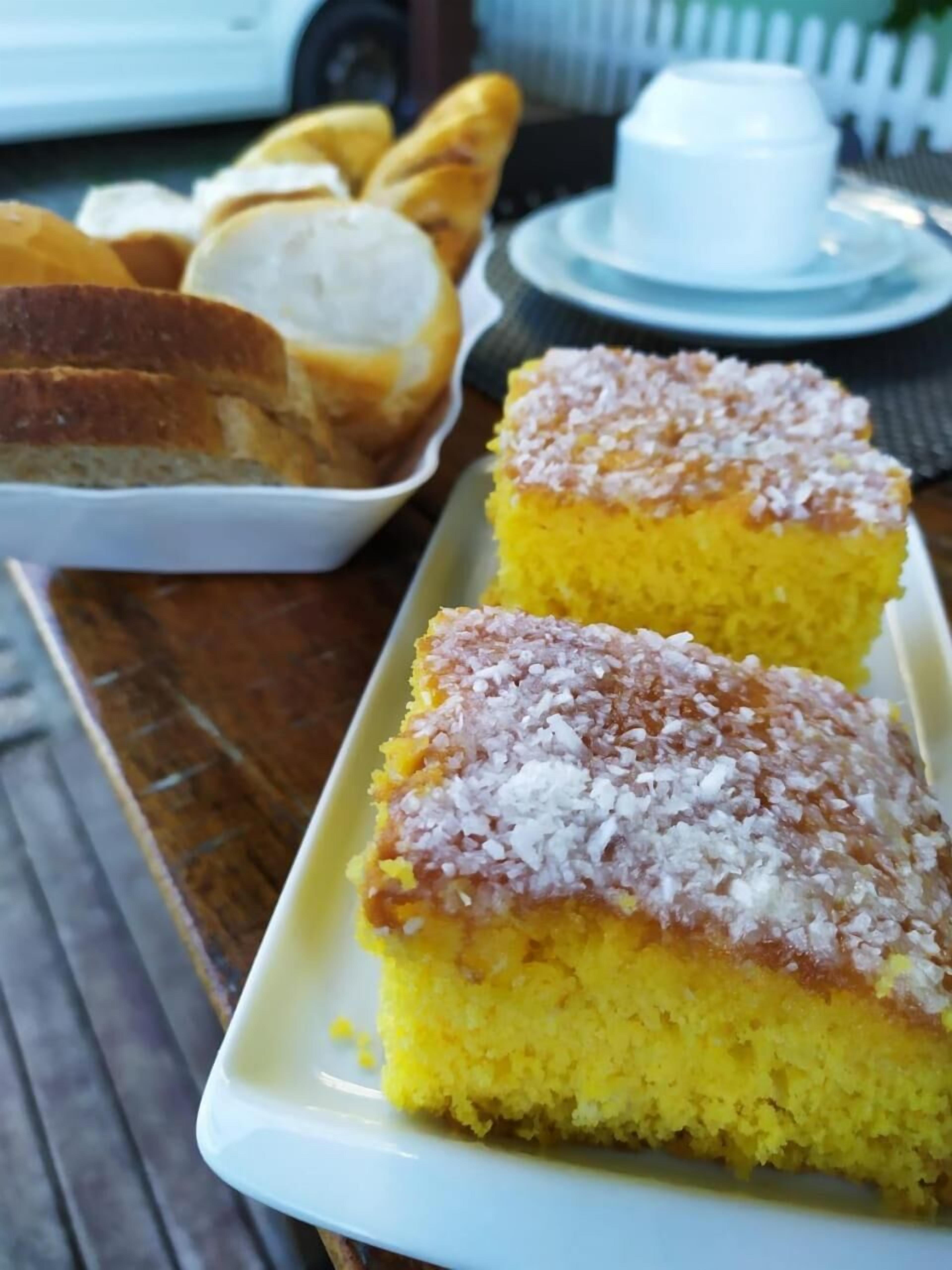 Un plateau sur lequel reposent deux carrés de gâteau jaune, recouverts de sucre blanc, une petite tasse blanche et plusieurs tranches de pain, le tout posé sur une table en bois.
