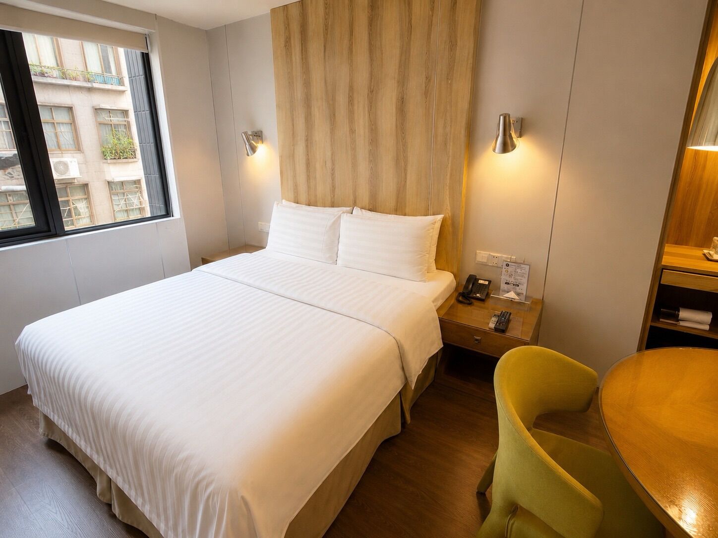 Habitación de hotel con cama, mesita de noche, lámpara, silla y vistas a edificios.