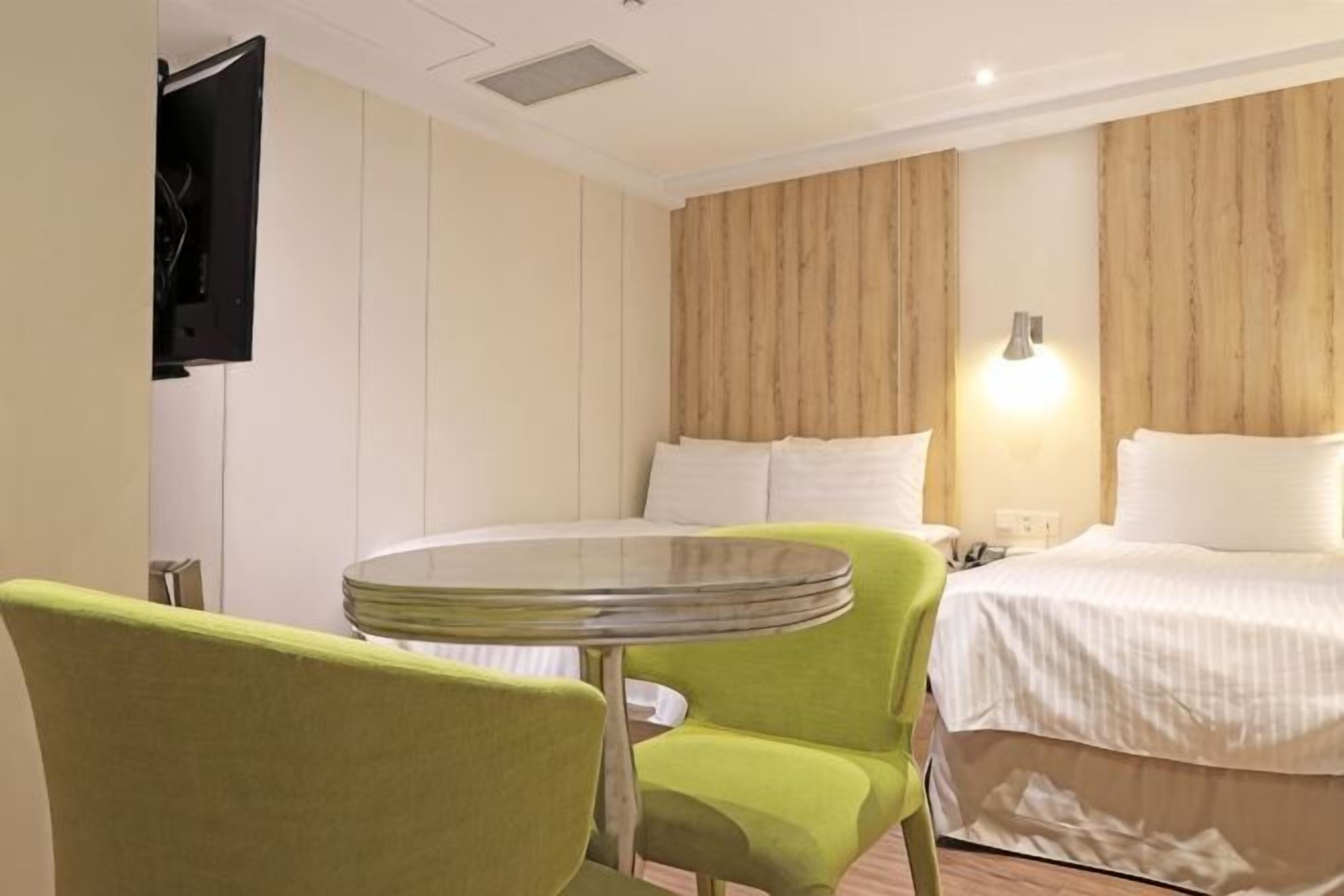 A hotel room with a bed, a round table, a green chair, a TV, and a wall-mounted lamp.