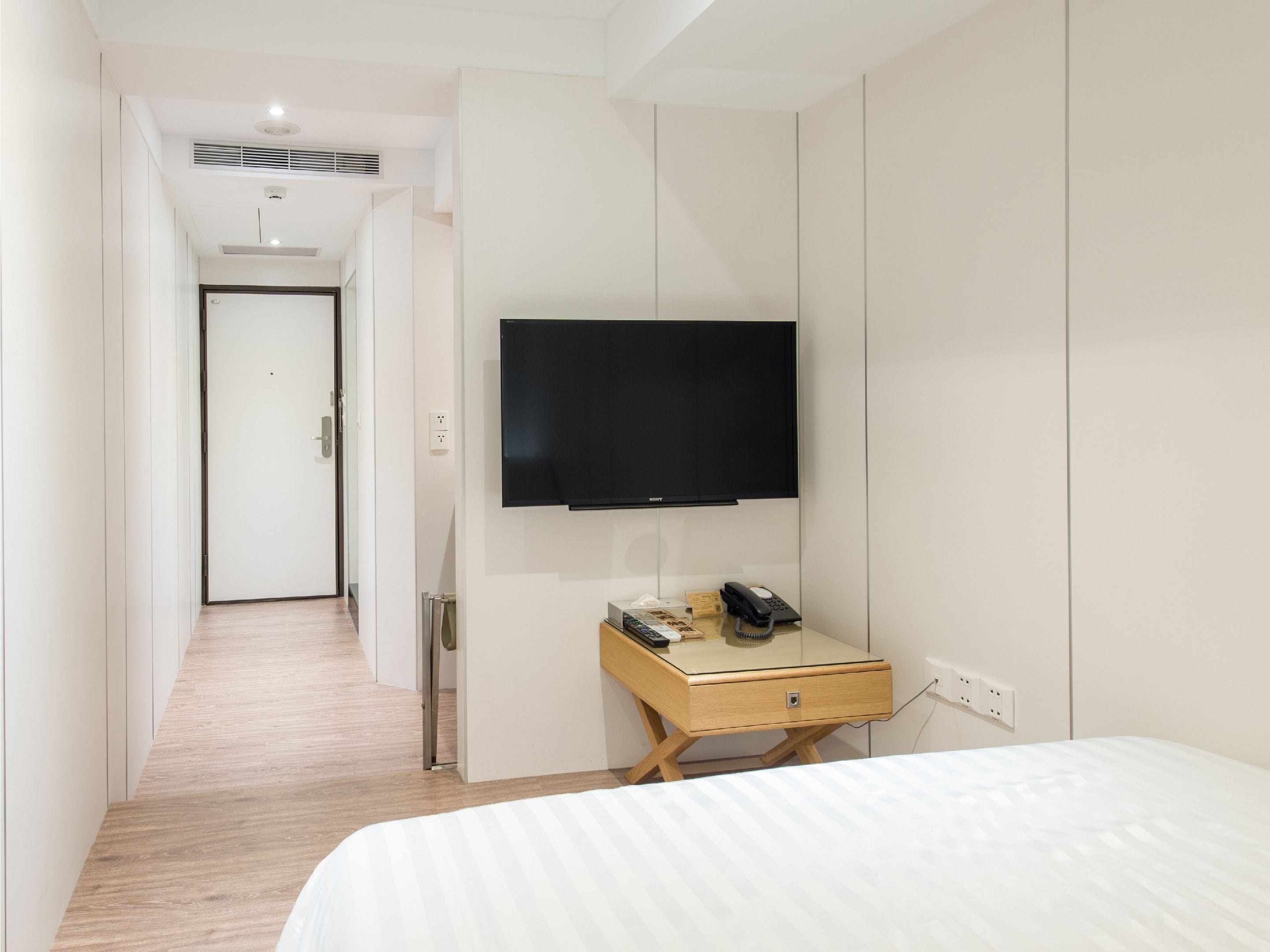 A hotel room with a bed, a desk with a telephone and a lamp, a flat-screen TV mounted on the wall, and a wooden floor.