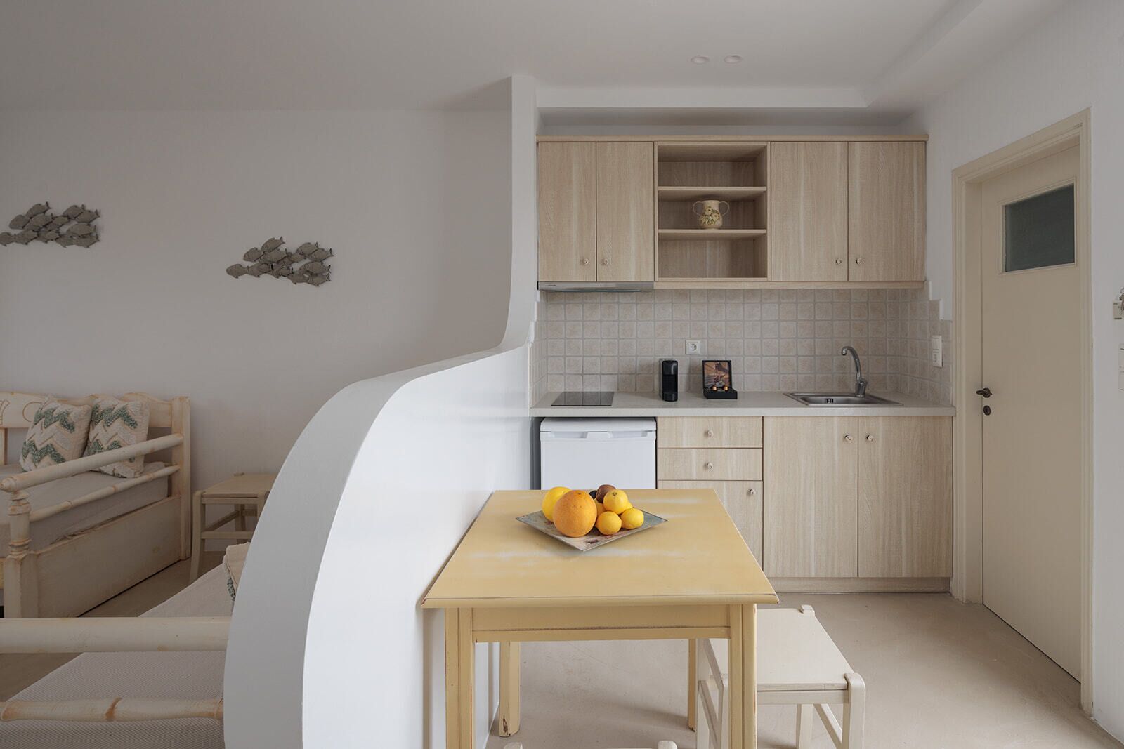 A compact kitchen with a dishwasher, a sink, and wooden cabinets. There is a small table with a fruit bowl.