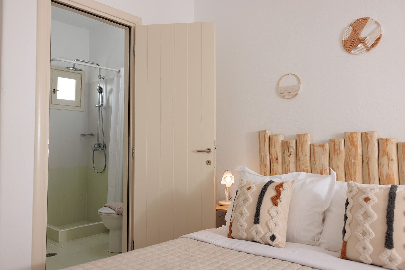 Un dormitorio con cama, cabecera de madera, mesita de noche con lámpara y zona de ducha visible a través de una puerta abierta.