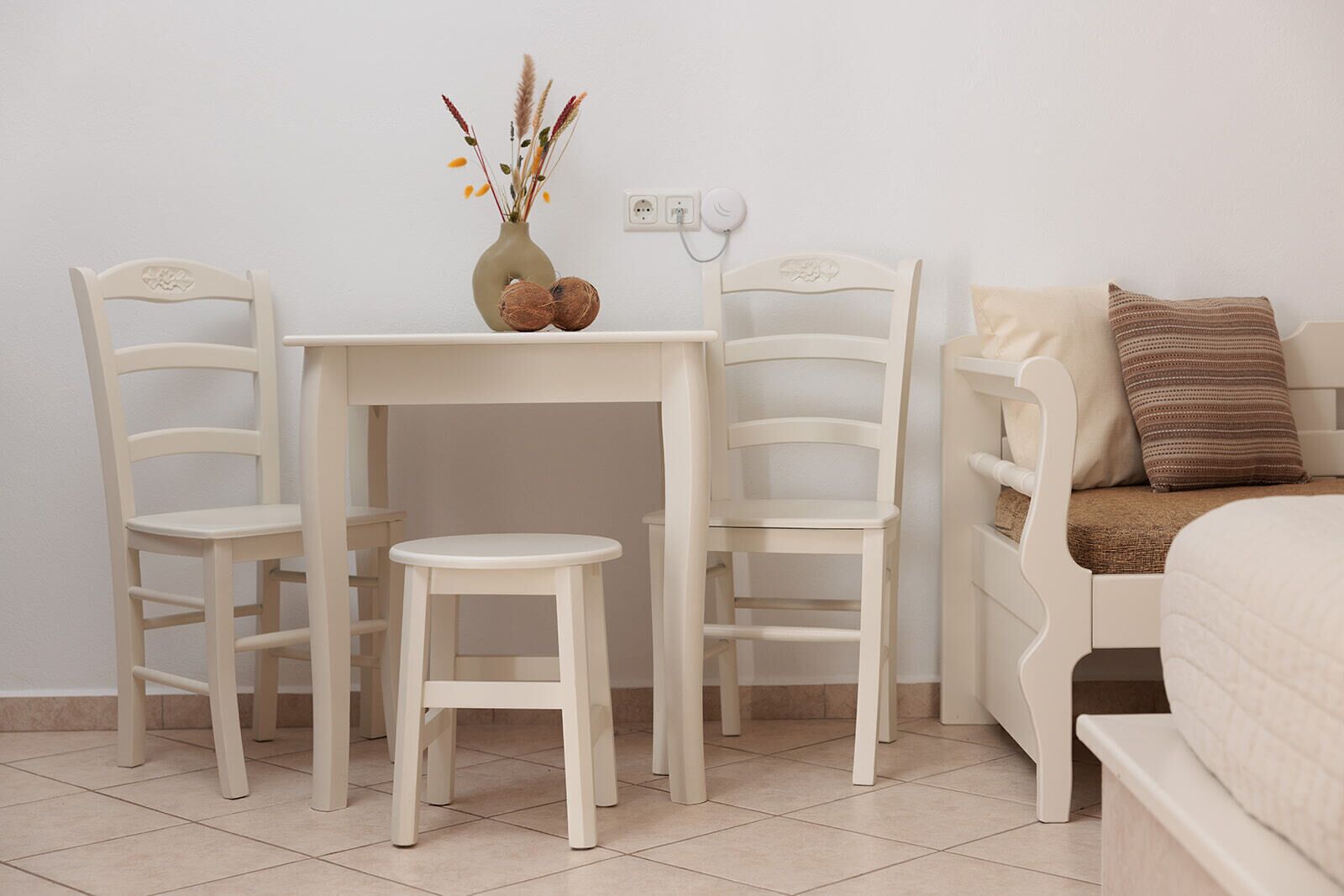 Une petite table blanche à deux places, sur laquelle est posé un vase contenant des plantes séchées, est flanquée de deux chaises blanches. Un lit avec un oreiller marron se trouve à droite.
