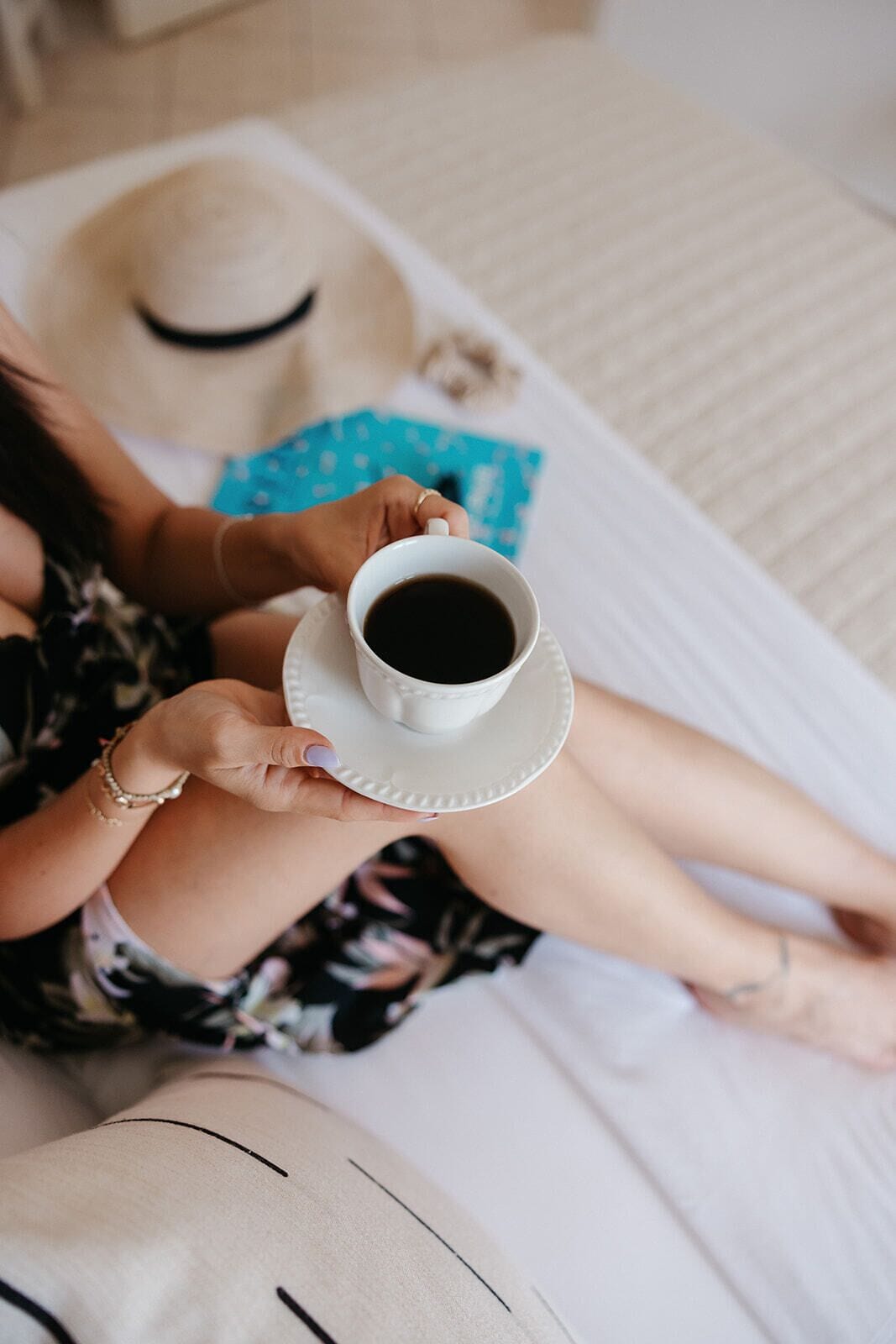 A person holding a cup of coffee on a white tray, lying on a bed with a hat and a blue book nearby.