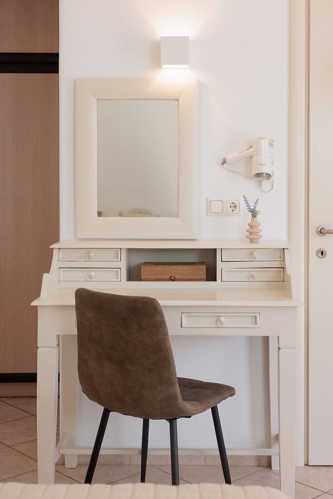 Un escritorio blanco con un espejo, un secador de pelo y un florero con flores.