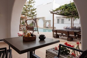 A patio area with a table, chairs, a wooden bowl with fruits, a small pot, and a pool in the background. - Hotel Anemomilos (Naxos)