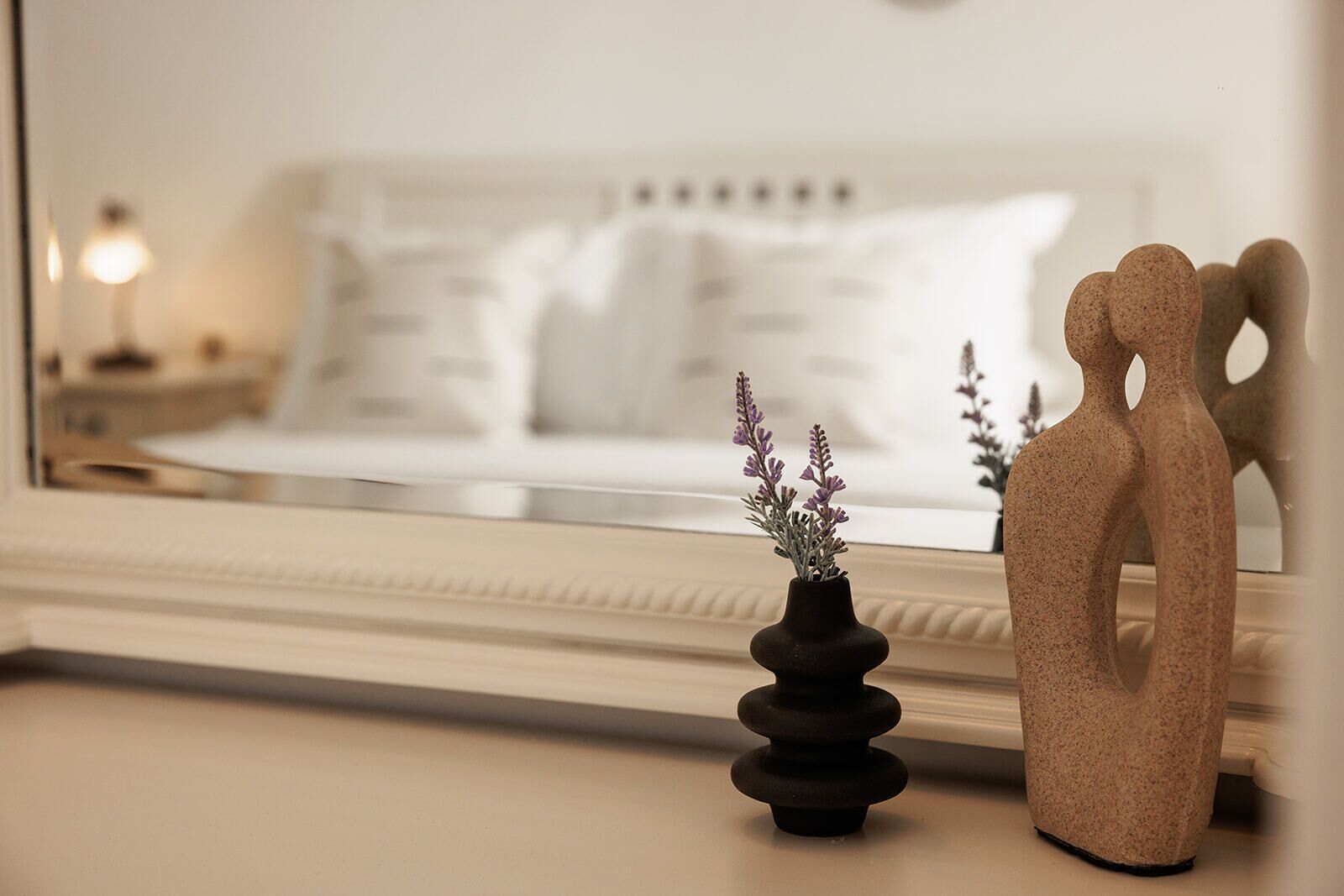 A bedroom with a bed, a nightstand, a lamp, and decorative items including a vase with flowers and a sculpture.