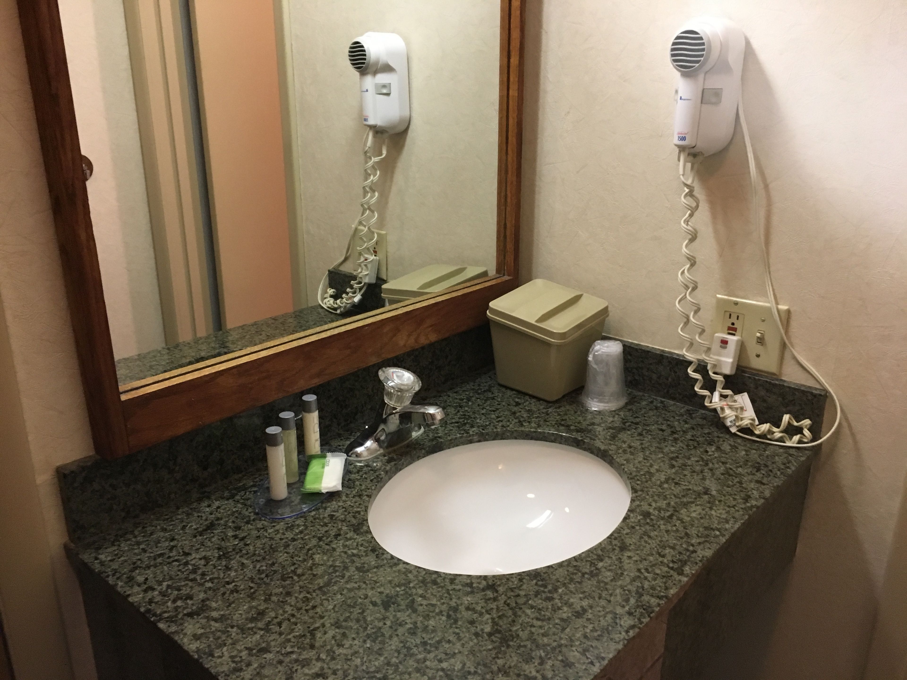 Un baño con encimera de granito, un lavamanos blanco, un espejo, dos secadores de pelo montados en la pared y un cubo de basura.