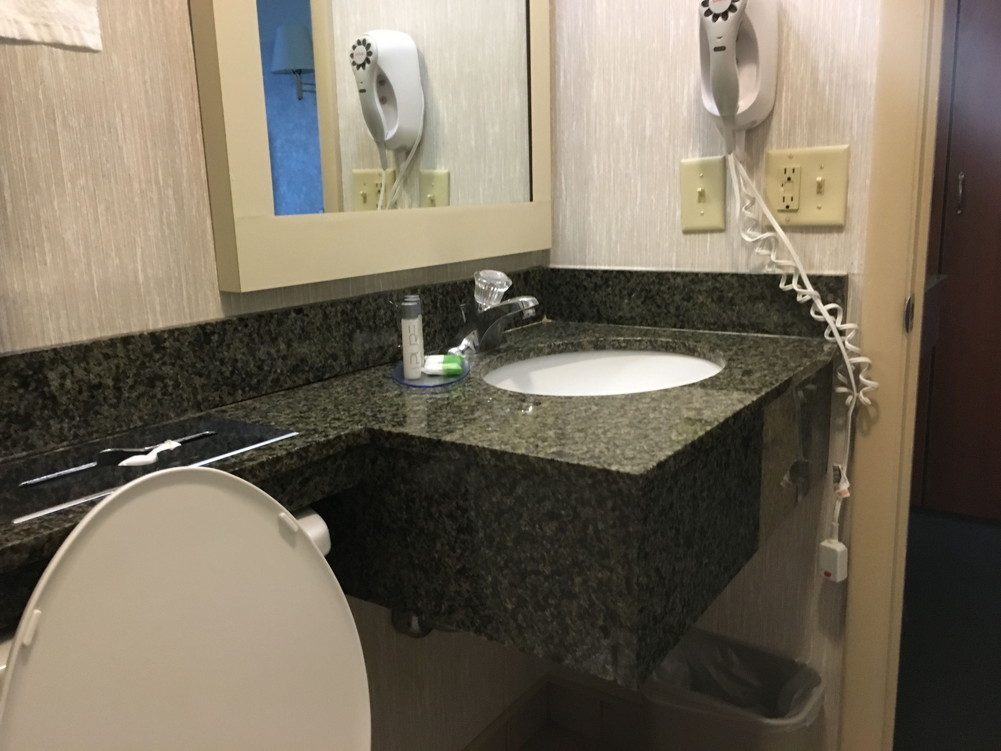 A bathroom with a granite countertop, a white sink, a mirror, a hairdryer, and a trash can.