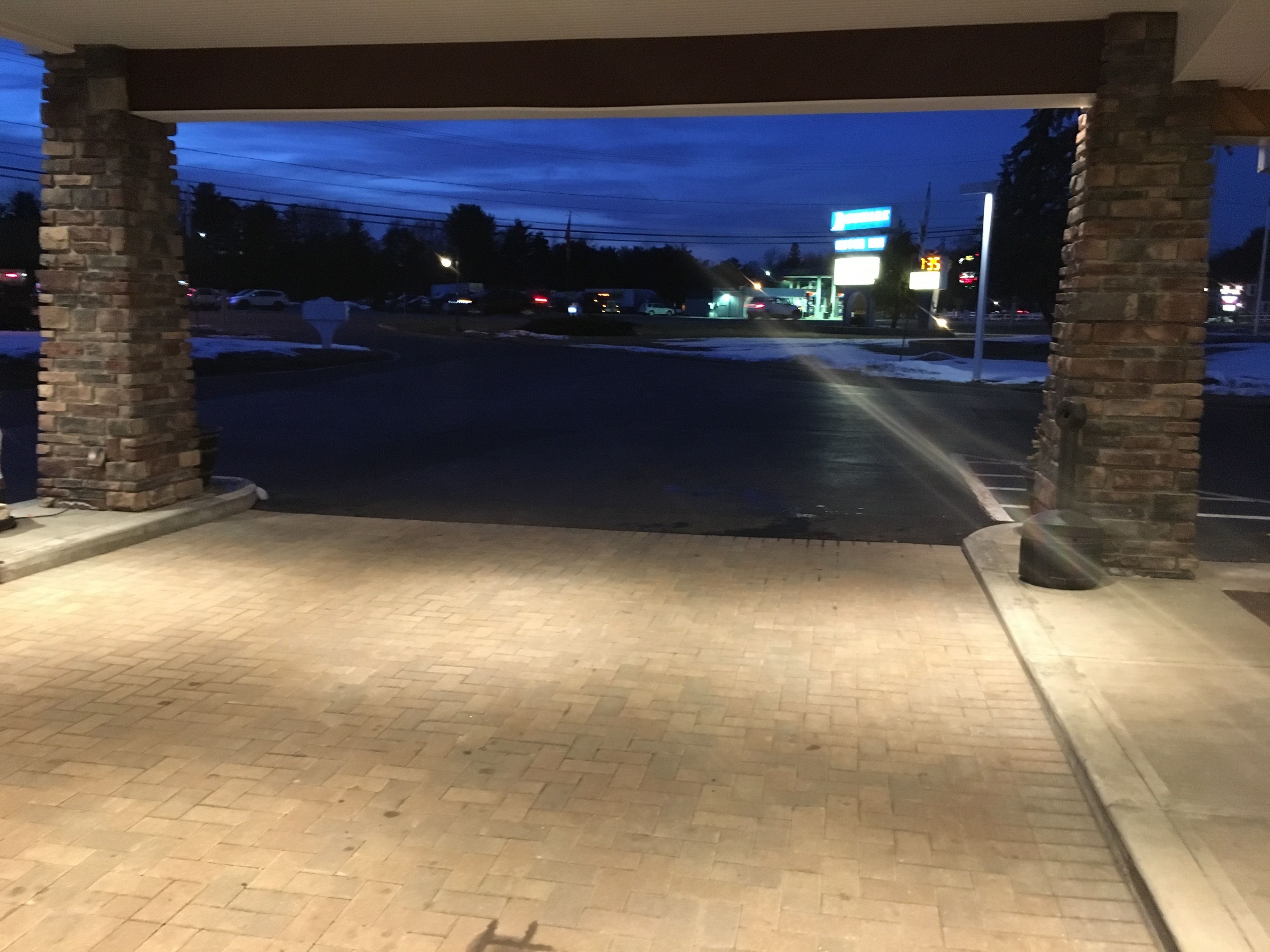 A covered outdoor area with brick flooring and stone pillars, leading to a parking lot with a gas station in the background.