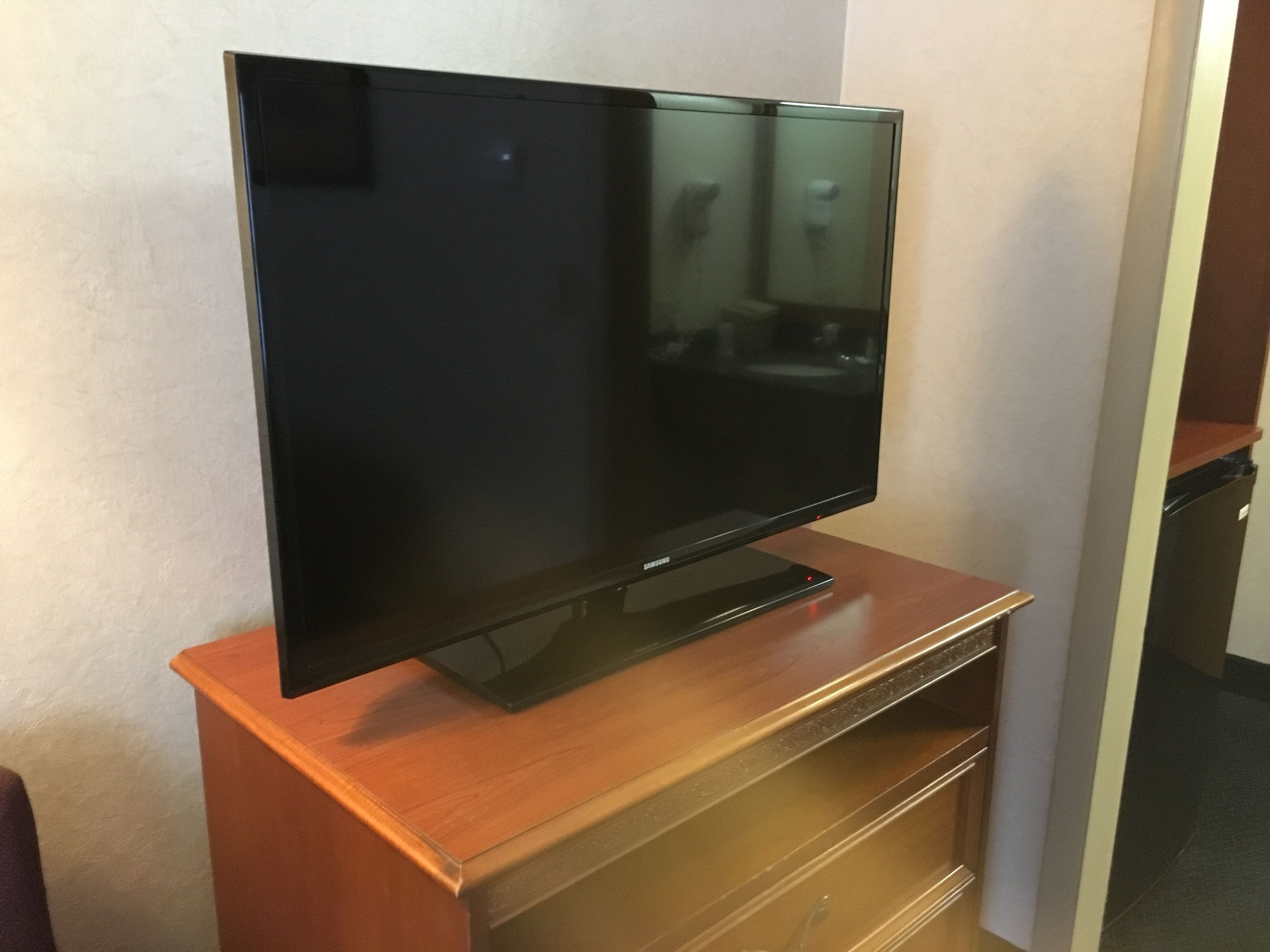 Un televisor de pantalla plana sobre un soporte de madera en una habitación con pared blanca y una puerta al fondo.