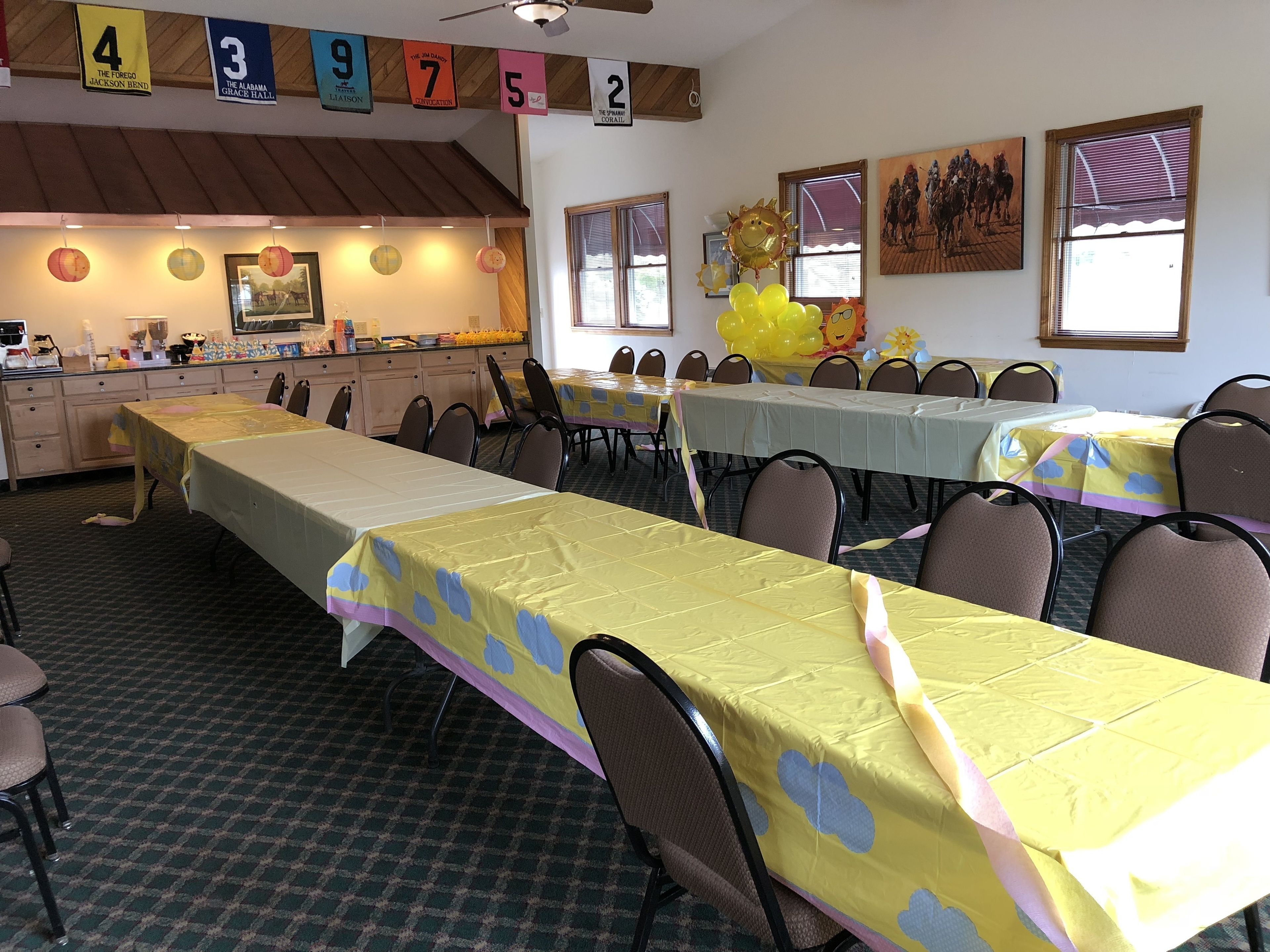 Un salón de banquetes con varias mesas cubiertas de manteles amarillos, sillas dispuestas alrededor y una zona de cocina al fondo.