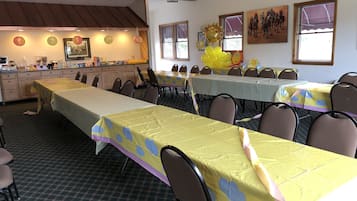 Un salón de banquetes con varias mesas cubiertas de manteles amarillos, sillas dispuestas alrededor y una zona de cocina al fondo.