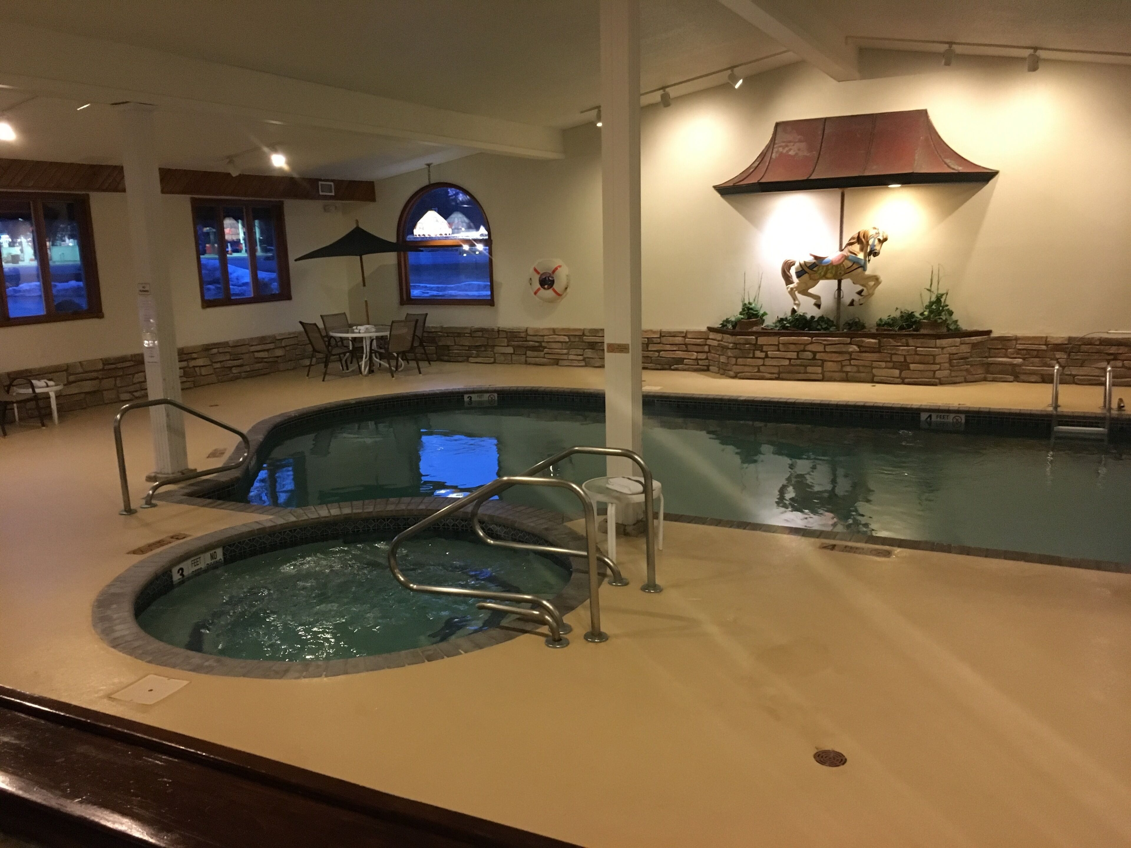An indoor swimming pool with a hot tub, stone walls, and a view of a building through a window.