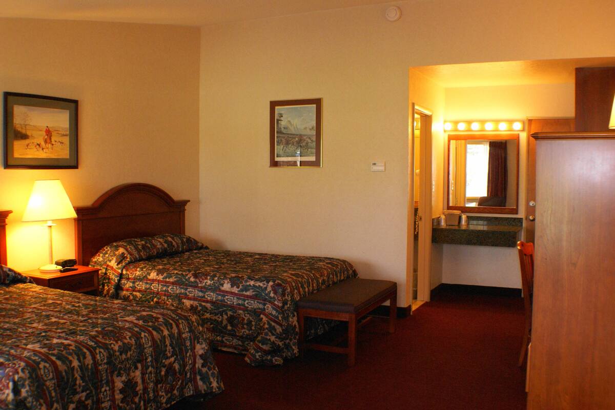 A hotel room with two beds, a bench, a mirror with lights, and a framed picture on the wall.