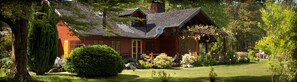 A rustic log cabin with a sloped roof, surrounded by lush greenery and a well-maintained garden. - Hotel Posada del Río (Pucón)