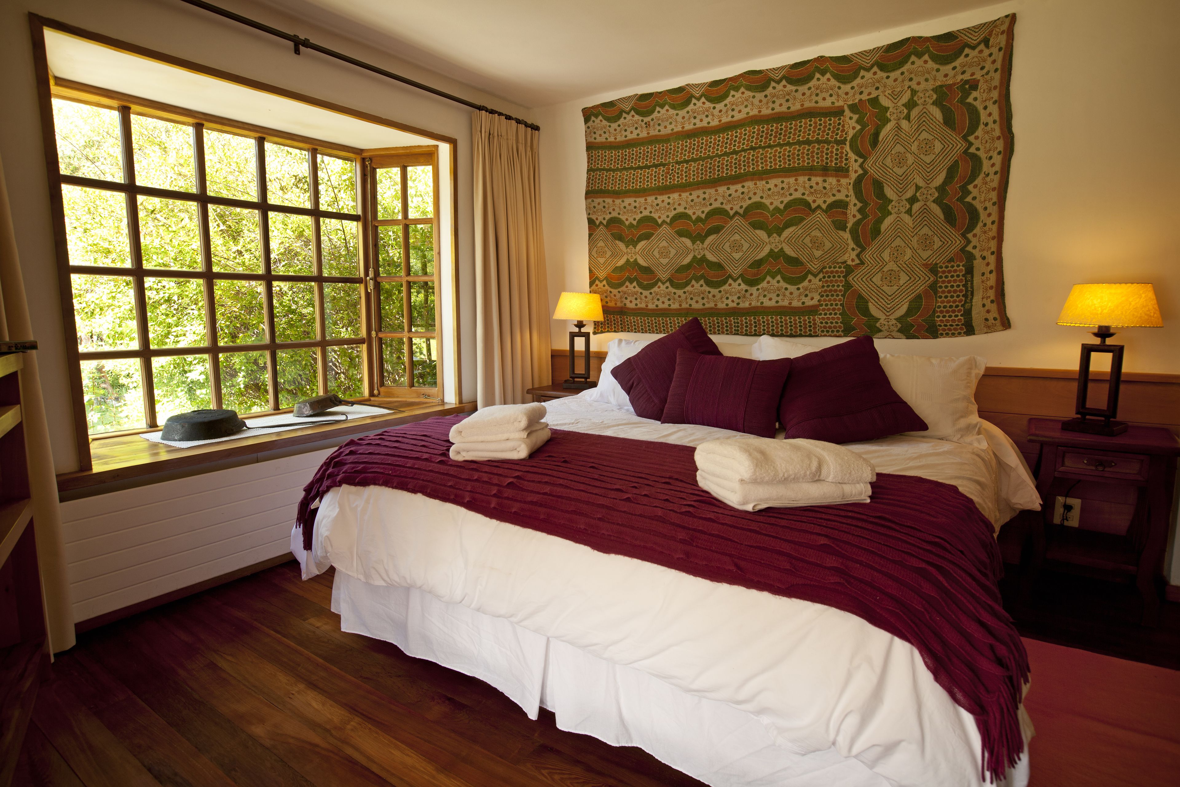A neatly made bed with a red and white bedspread, two bedside lamps, and a large window with curtains.