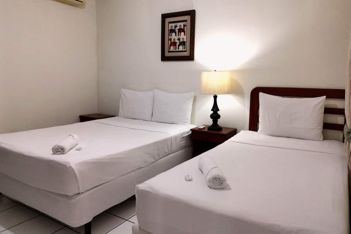 Habitación de hotel con dos camas individuales, ropa de cama blanca, una mesita de noche con lámpara, un cuadro en la pared y un equipo de aire acondicionado.