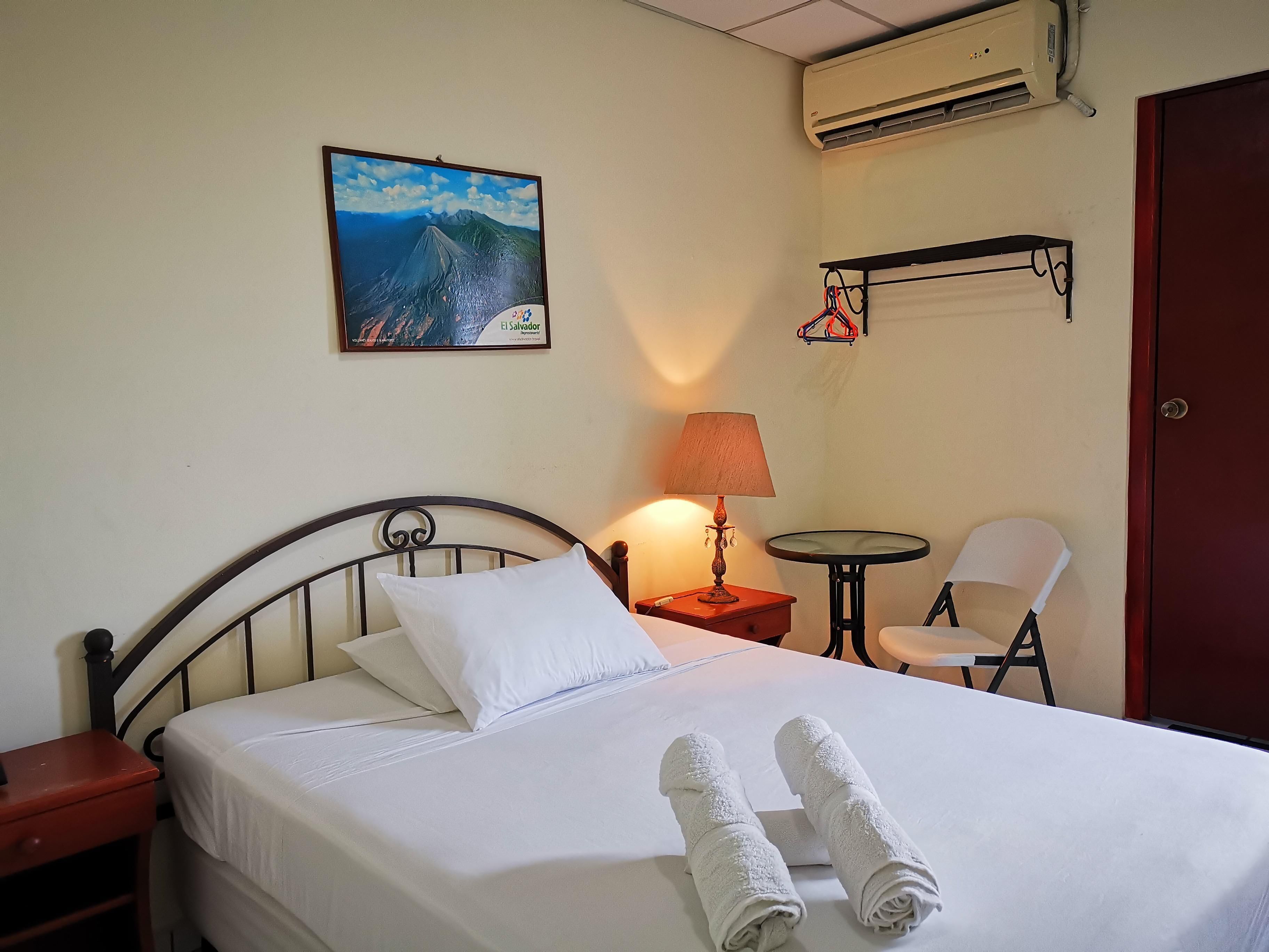 Habitación de hotel con cama, mesita de noche, lámpara, silla, pequeña mesa, equipo de aire acondicionado y un cuadro en la pared.