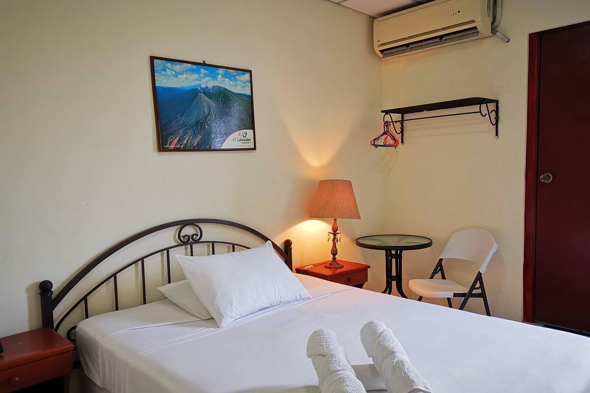Habitación de hotel con cama, mesita de noche, lámpara, silla, pequeña mesa, equipo de aire acondicionado y un cuadro en la pared.