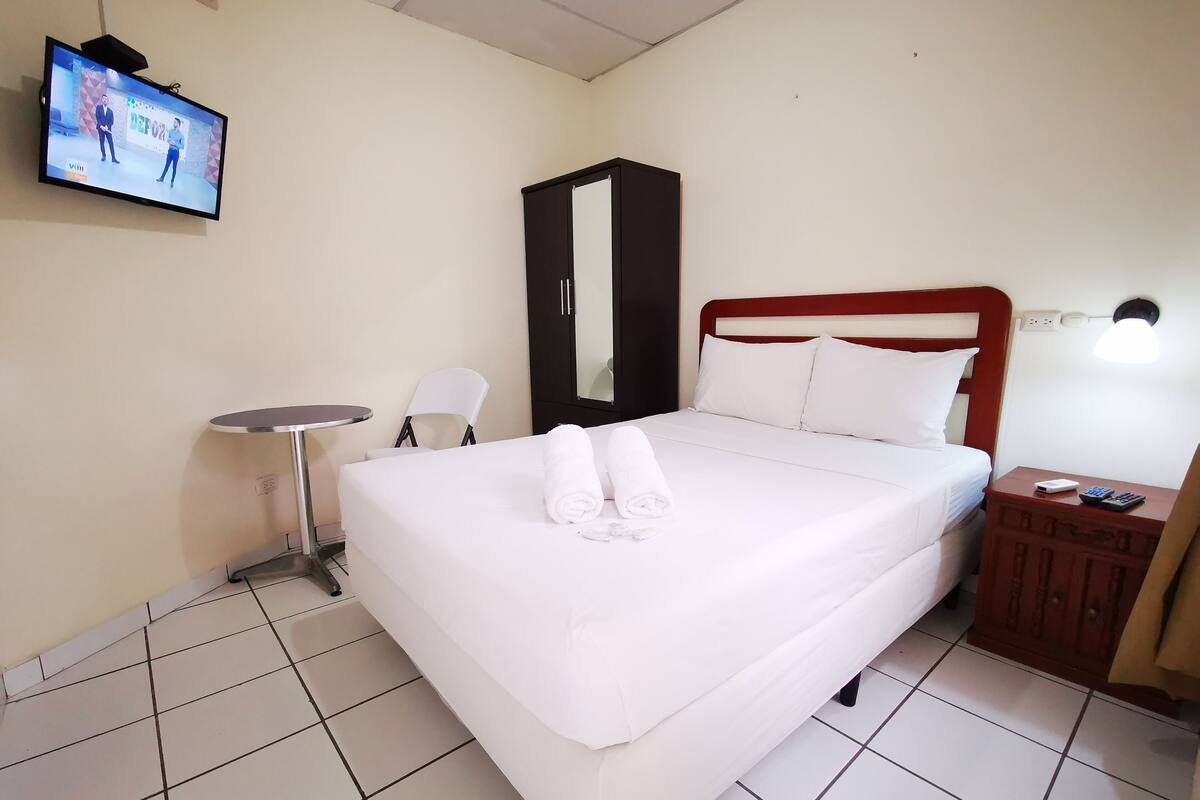 Una habitación de hotel con cama, televisor montado en la pared, una mesita, una silla y un armario.
