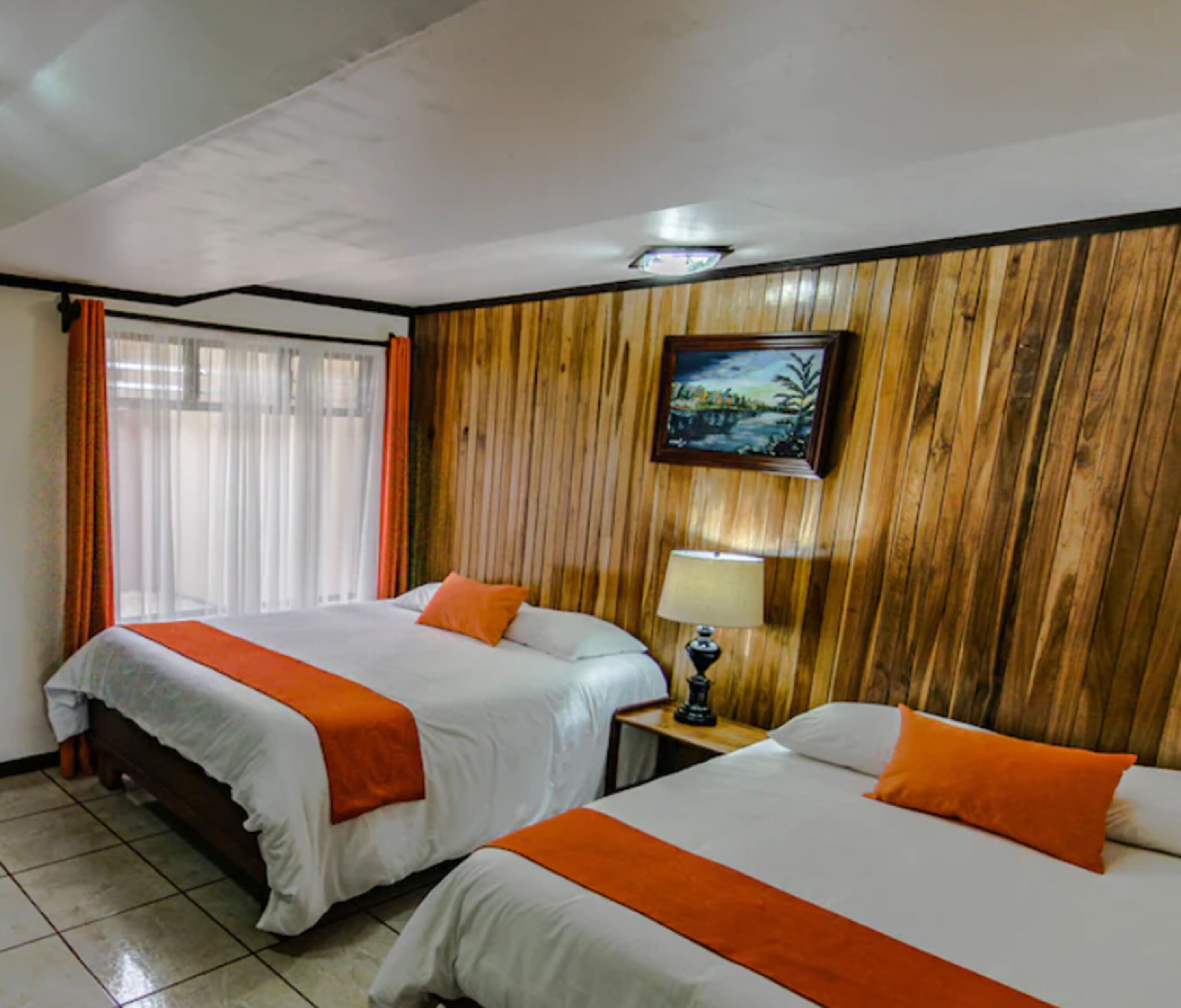 A hotel room with two beds, wooden paneling on the walls, a painting, and a lamp on a bedside table.