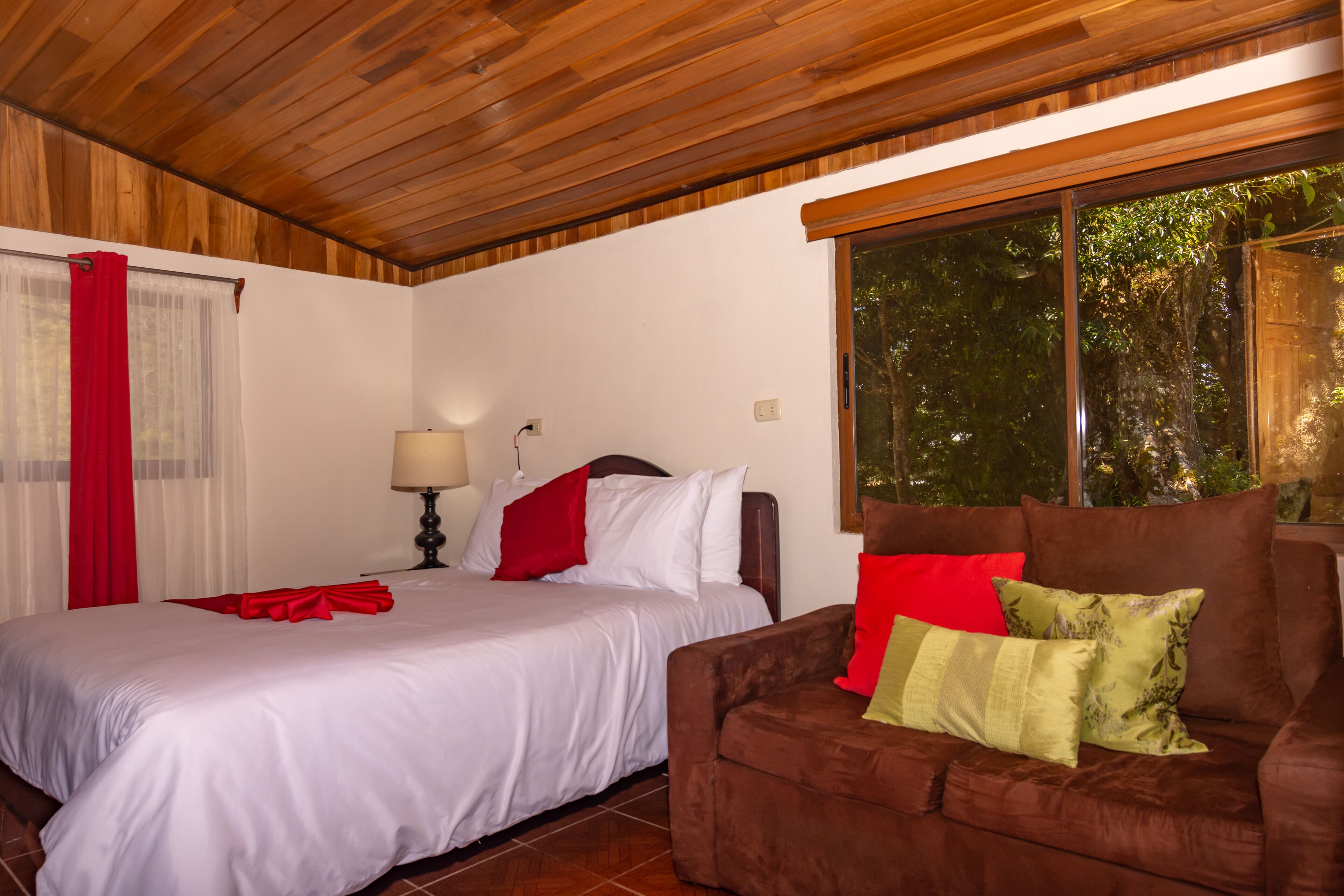Une chambre à coucher avec un lit, un canapé marron, une fenêtre donnant sur des arbres et une lampe.