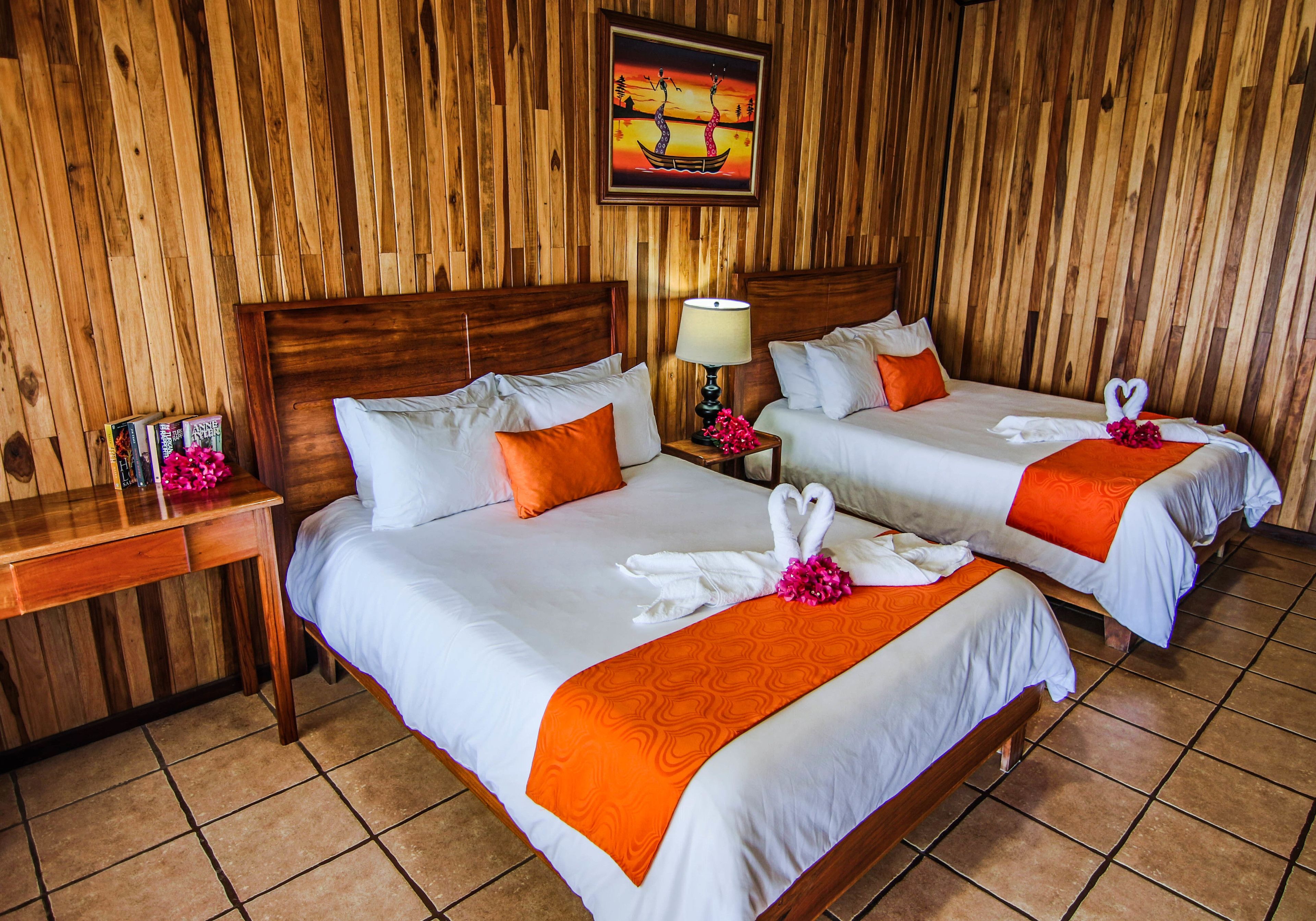 A hotel room with two beds, wooden walls, a nightstand with books and a lamp, and a painting on the wall.