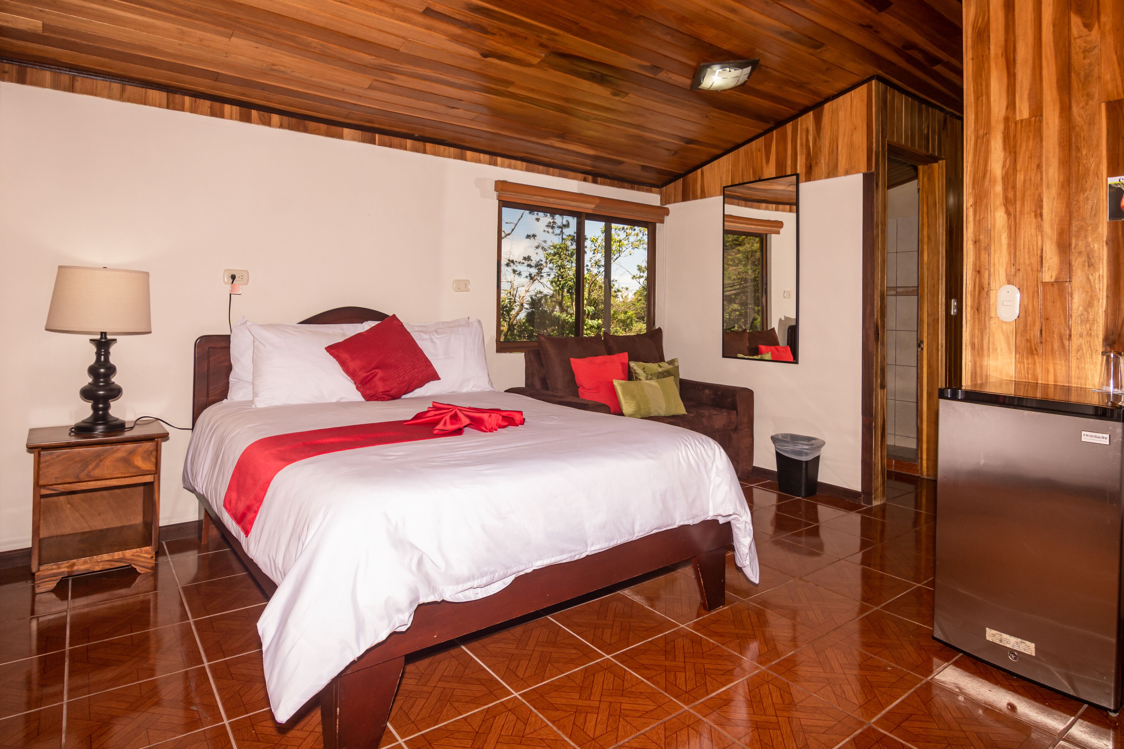 A hotel room with a wooden ceiling, a bed with white bedding and red accents, a nightstand with a lamp, a small trash bin, and a refrigerator.
