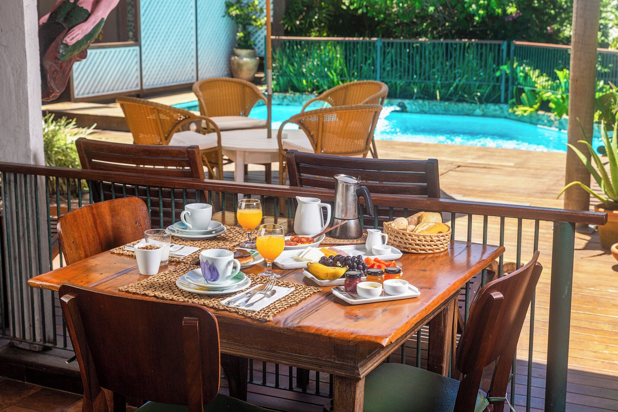 Mesa de madera con elementos de desayuno, rodeada de sillas de mimbre, con vista a una piscina.