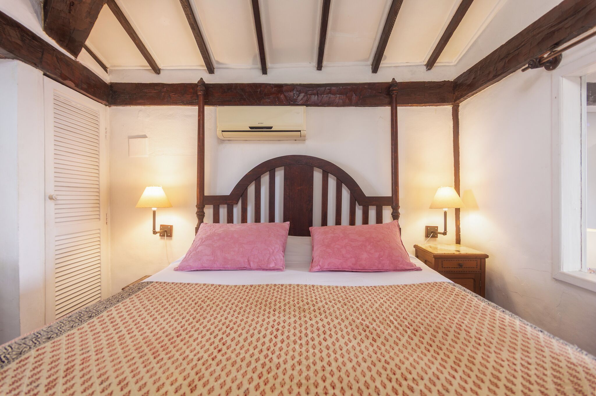 A bedroom with a bed, two bedside lamps, a nightstand, a window, and an air conditioning unit.
